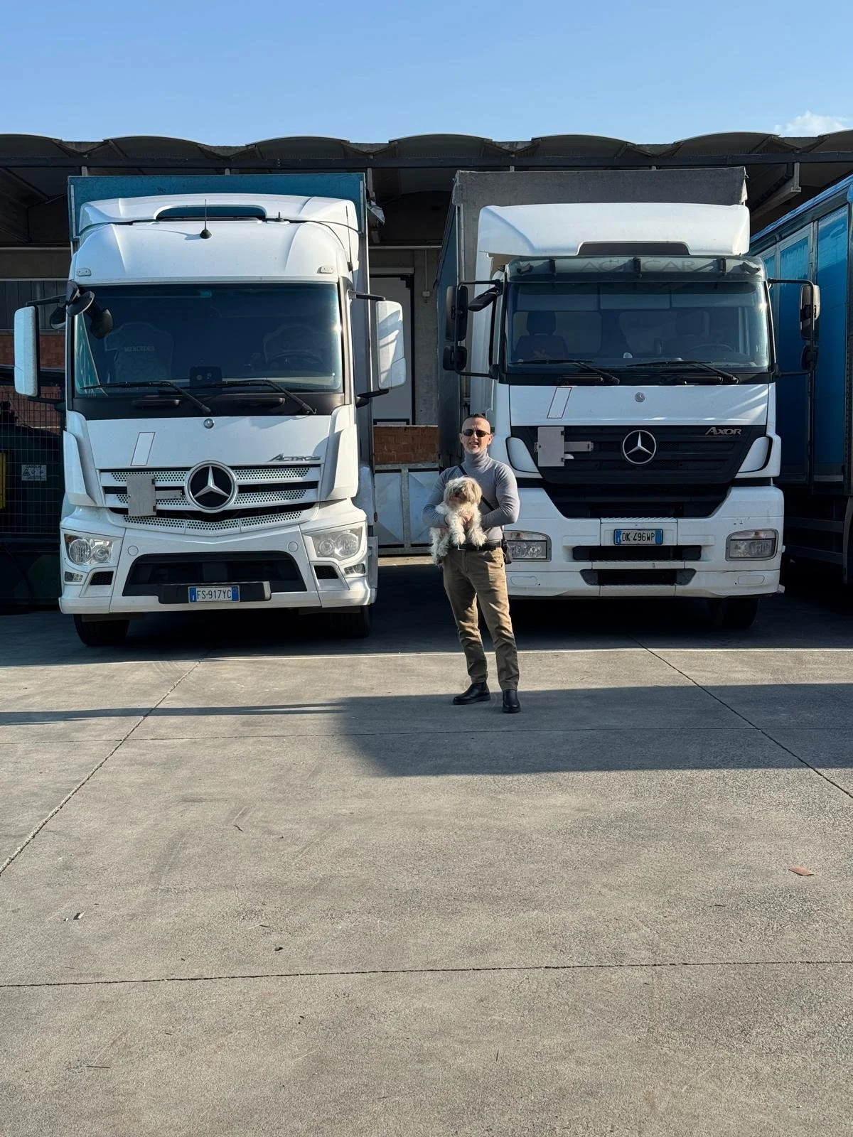 Un uomo con occhiali che tiene in braccio un cane vicino a due camion Mercedes àrmadio, in un'area di parcheggio.