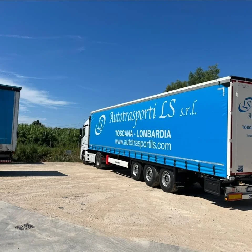 Un camion con scatola blu con scritte 'Autotrasporti LS s.r.l., Toscana - Lombardia, www.autotrasportils.com' parcheggiato su un terreno sterrato, sotto un cielo azzurro con alcune nuvole leggere.