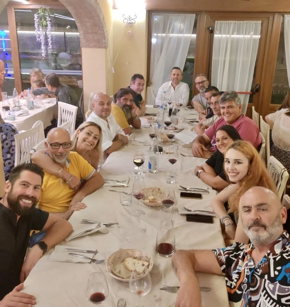 Un gruppo di persone sedute attorno a un grande tavolo durante una cena in un ristorante, con bicchieri di vino rosso e cibo sulla tavola.