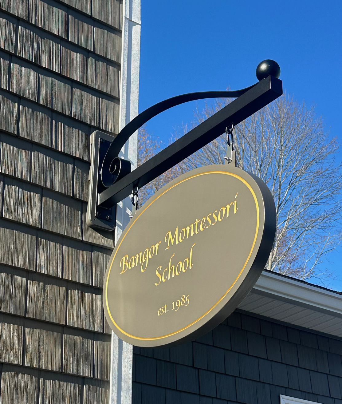 Sign for Bangor Montessori School hanging on a metal bracket outside, with a background of a blue sky and leafless tree.