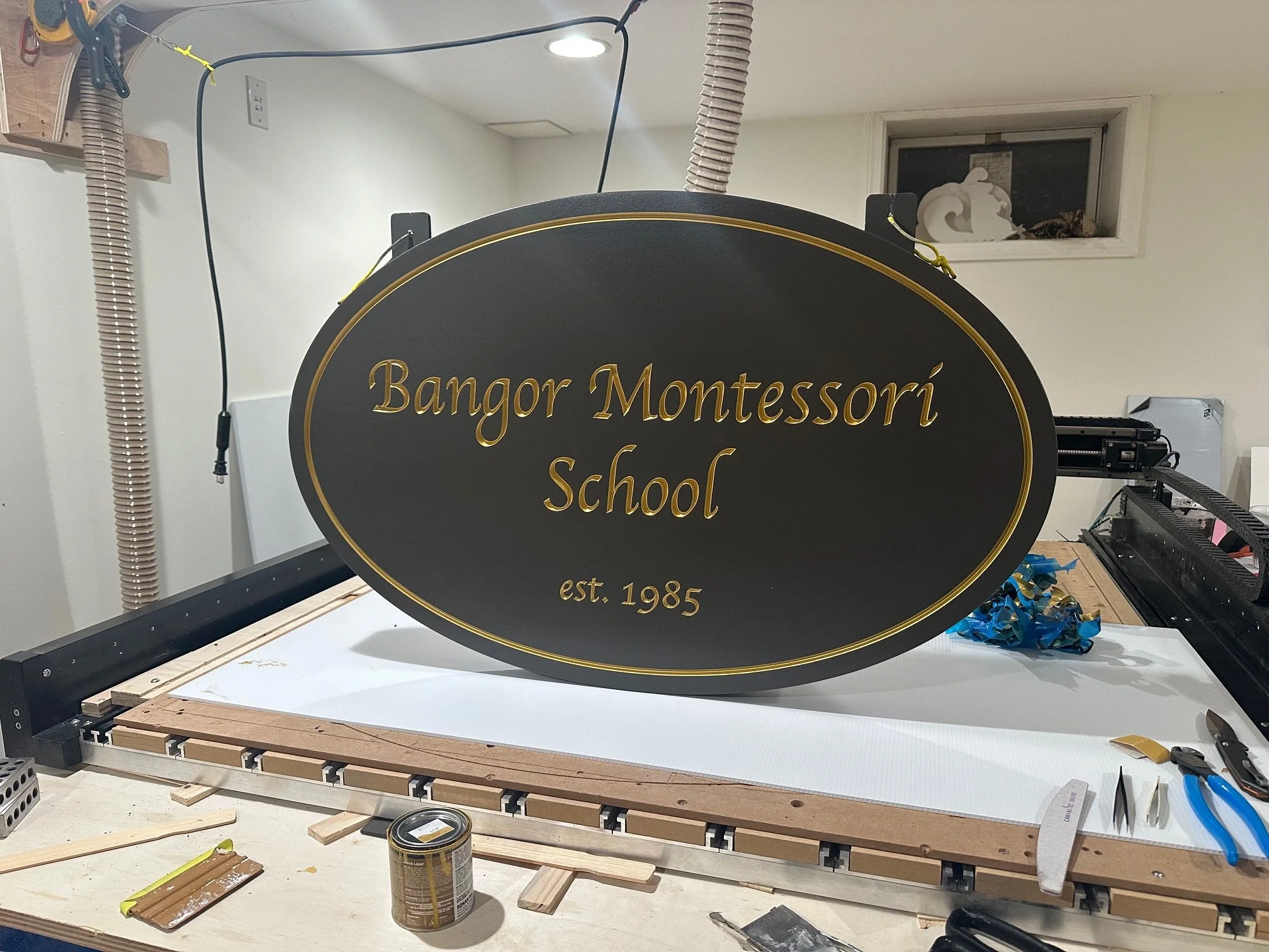 Black oval sign with gold lettering that reads "Bangor Montessori School est. 1985" on a worktable with tools and materials