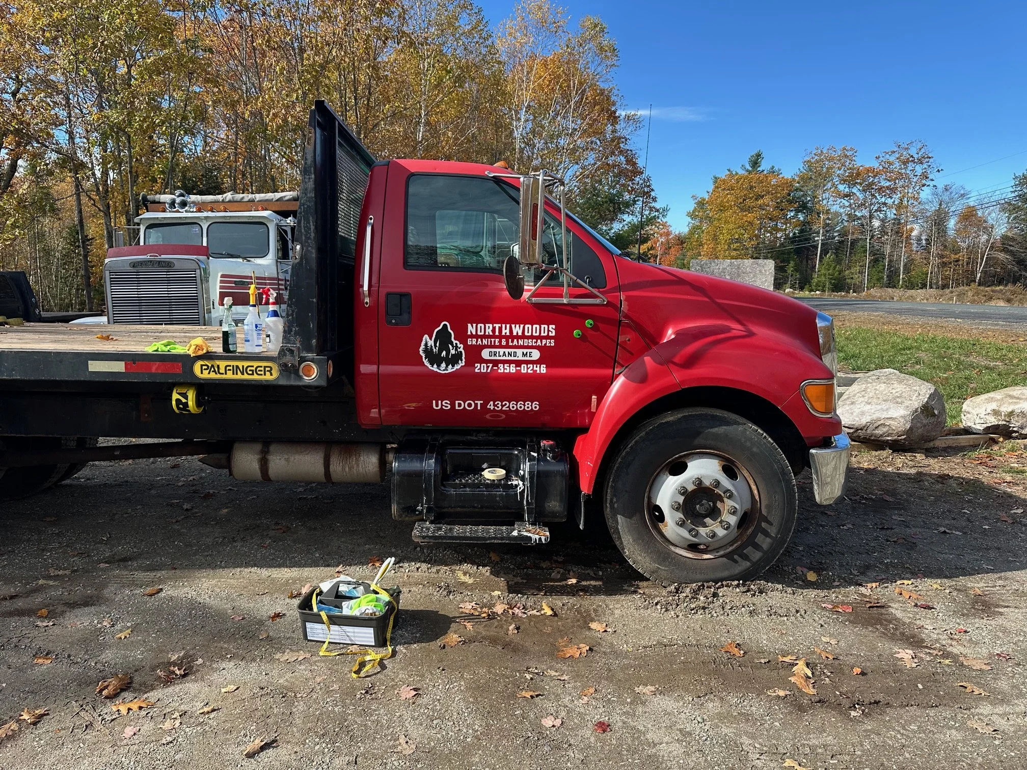 Red at work truck with company branding parked on a dirt surface with autumn trees in the background, a toolbox with tools and supplies on the ground nearby, and a similar vehicle on a flatbed trailer.