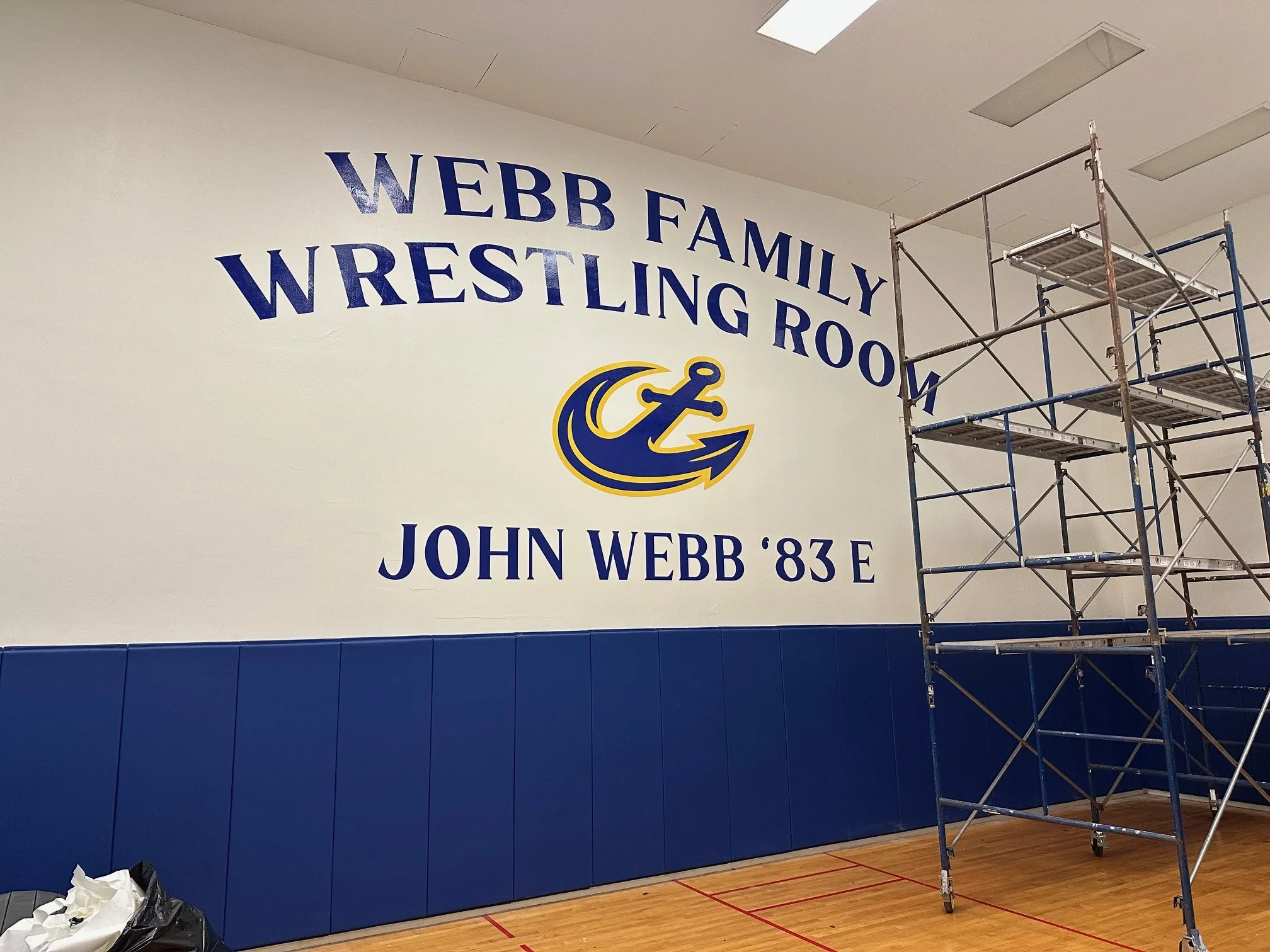 Wall decoration in the Webb Family Wrestling Room with text and logo. The text reads 'Webb Family Wrestling Room John Webb '83 E'. There is a logo featuring an anchor.  A scaffolding structure is also visible on the right side of the image.