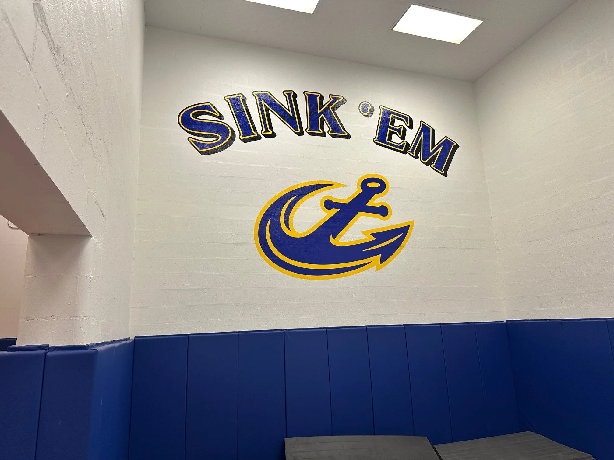 Interior of a room with white cinder block walls, featuring a large sign that reads "SINK EMS" in yellow and blue lettering, accompanied by a graphic of an anchor and wave symbol in matching colors, and blue padded benches below.