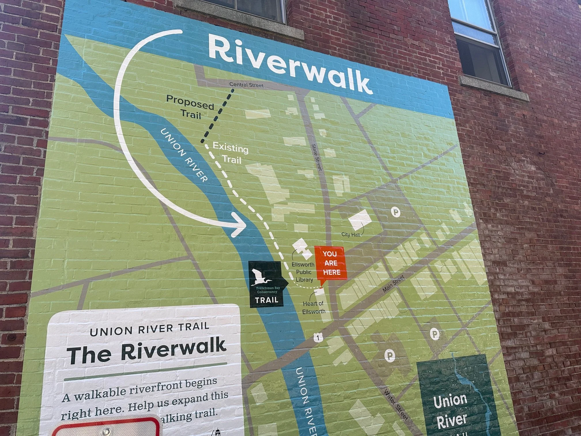 Map of the Union River Trail and Riverwalk in Ellsworth, Maine, showing the trail along the Union River, existing and proposed trails, the waterway, parking areas, the Ellsworth Public Library, City Hall, and a marked location with a red sign indicat