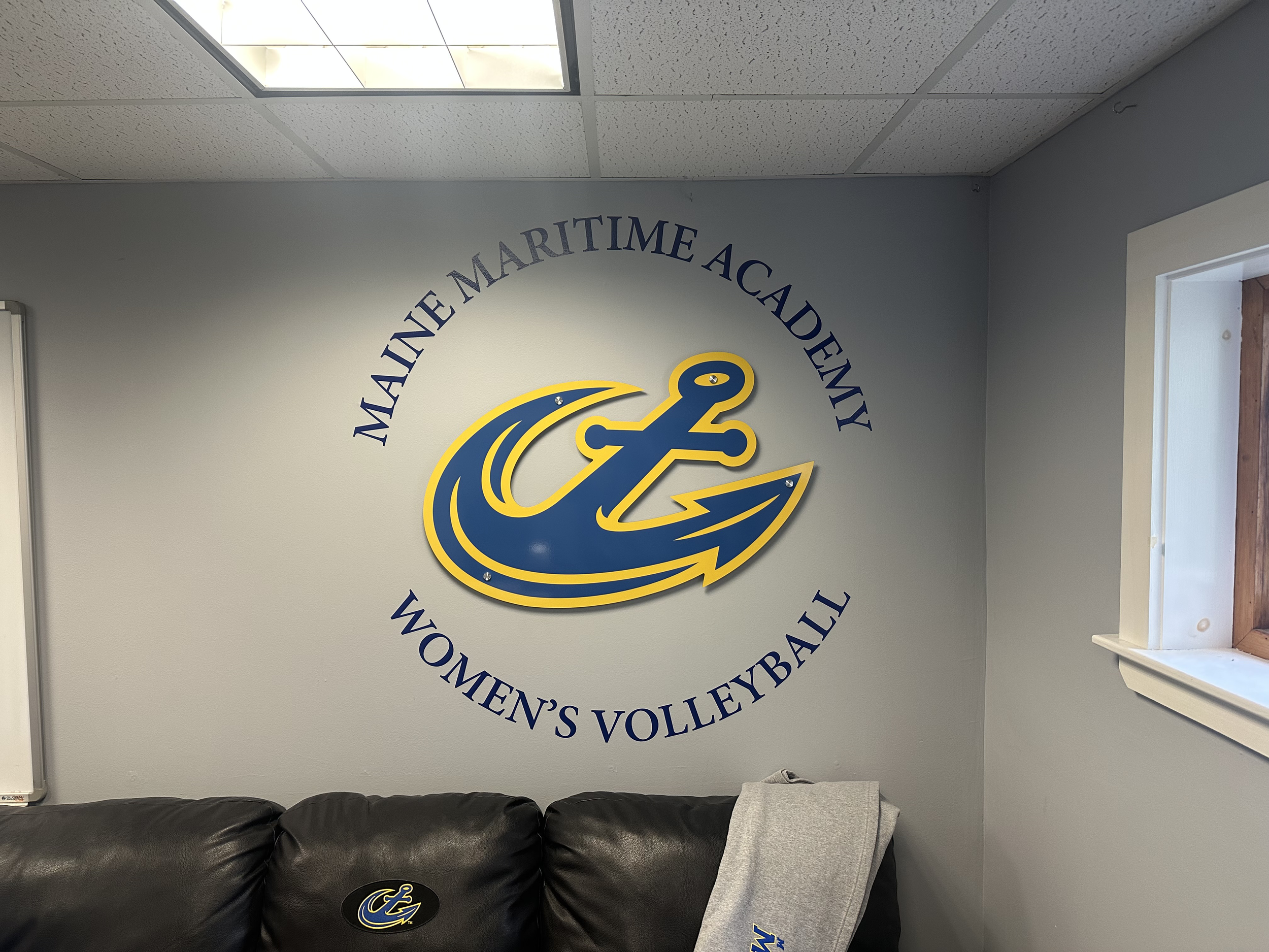 Wall decal displaying the logo for Maine Maritime Academy Women's Volleyball with a stylized anchor and arrow in blue and yellow, on a light-colored wall next to a window and a leather sofa.