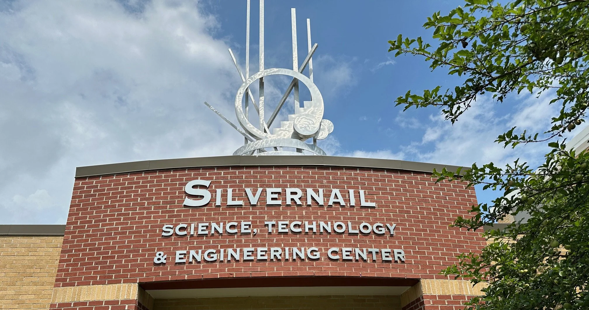 Sign for Silvernail Science, Technology & Engineering Center with sculpture on top, blue sky with clouds, and green tree branches to the right.