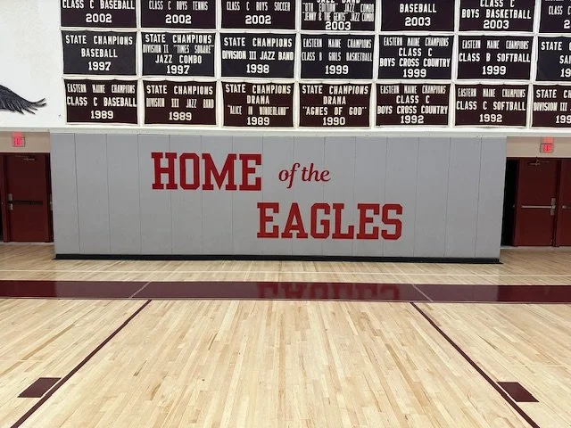 A gymnasium wall with a large sign that says "Home of the Eagles" in red letters. Above the sign are black plaques listing various sports achievements and championships. The gym floor is visible at the bottom of the image.