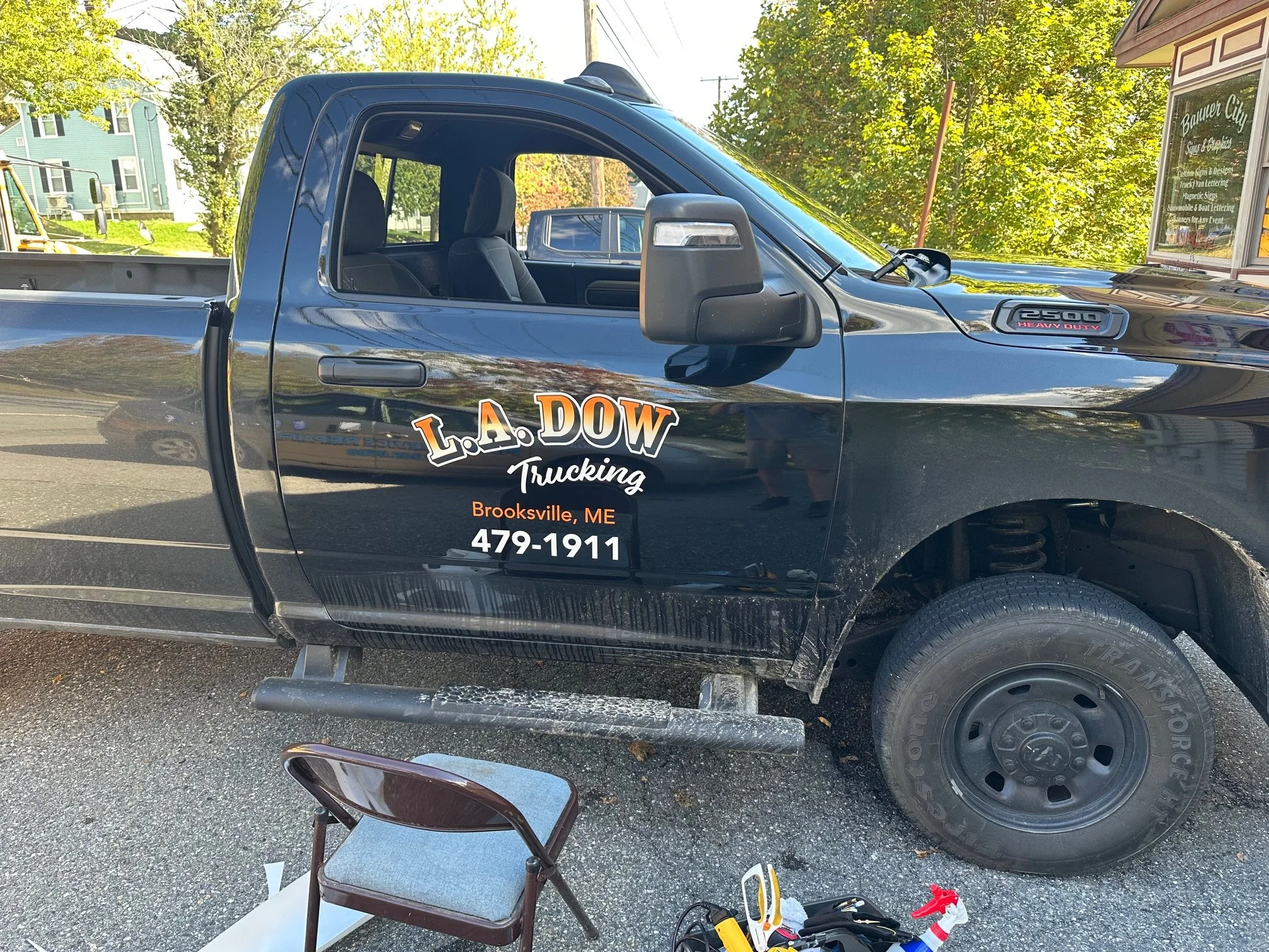 Black pickup truck with company branding on the door, parked outdoors, with a chair and tools on the ground nearby.