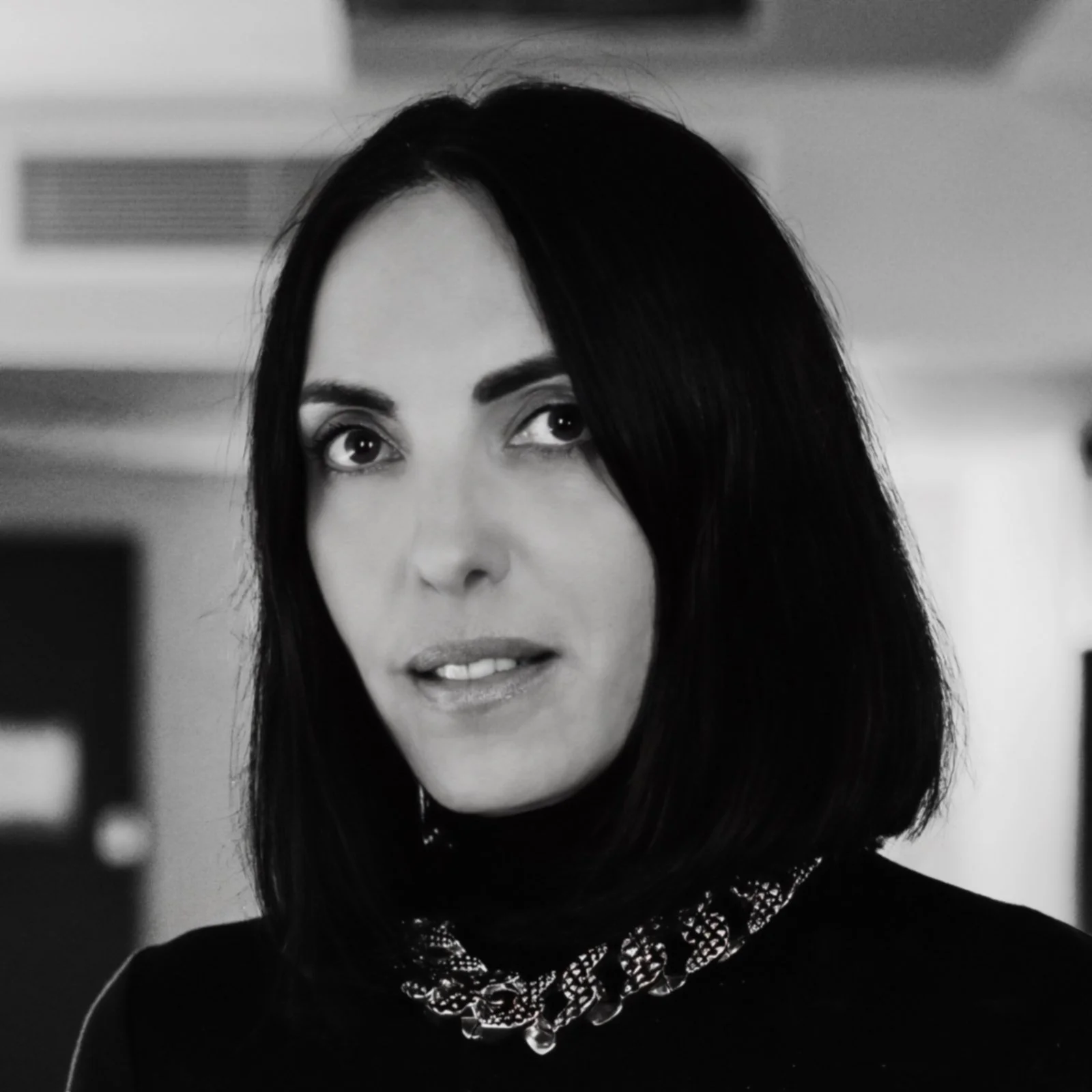 Black and white portrait of a woman with medium-length dark hair, wearing a patterned necklace, looking directly at the camera in an indoor setting.