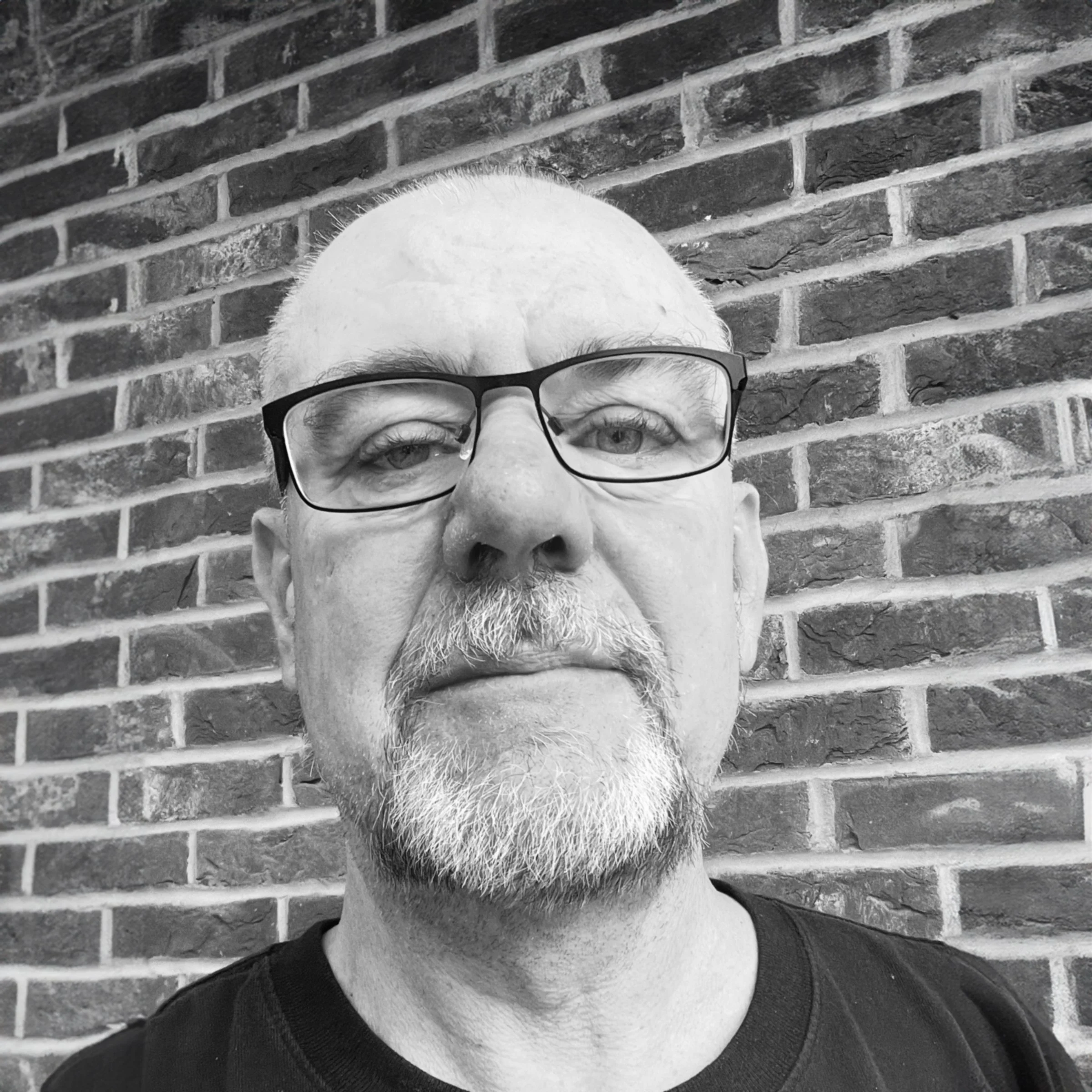 Black and white photo of an older man with glasses and a beard standing in front of a brick wall.