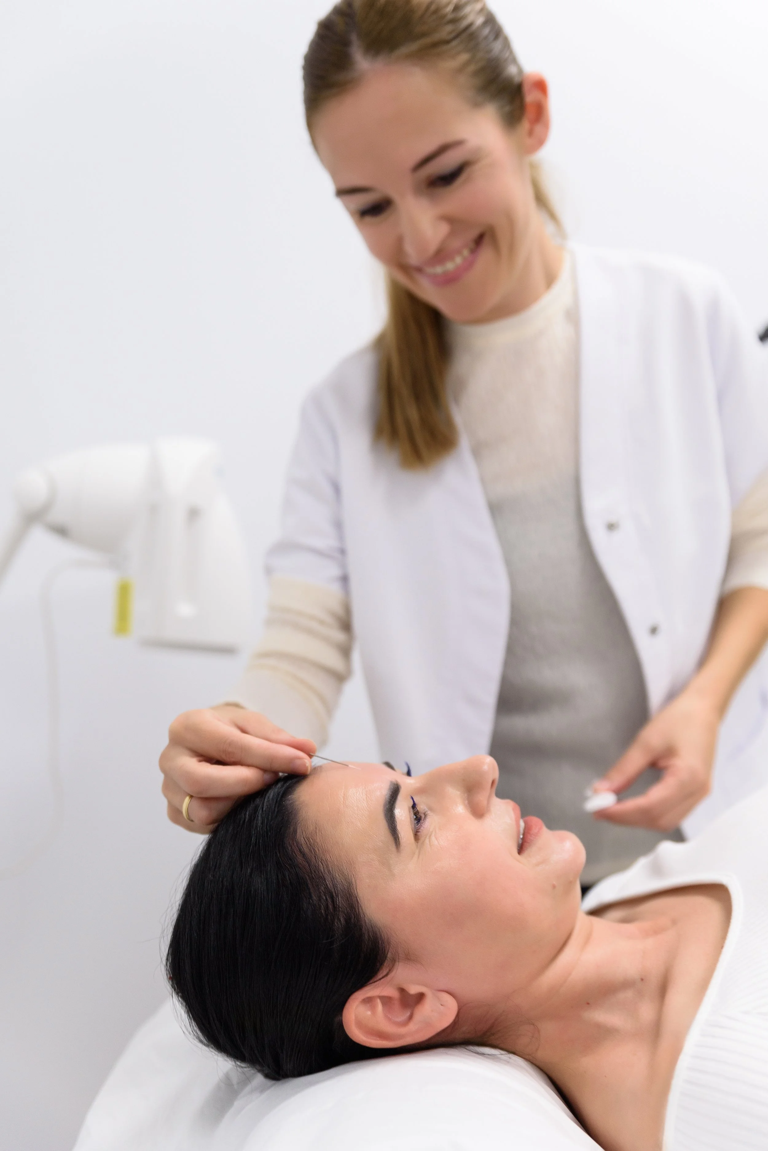 A woman lying down receiving a cosmetic treatment, with a professional woman using a small tool on her eyebrow.
