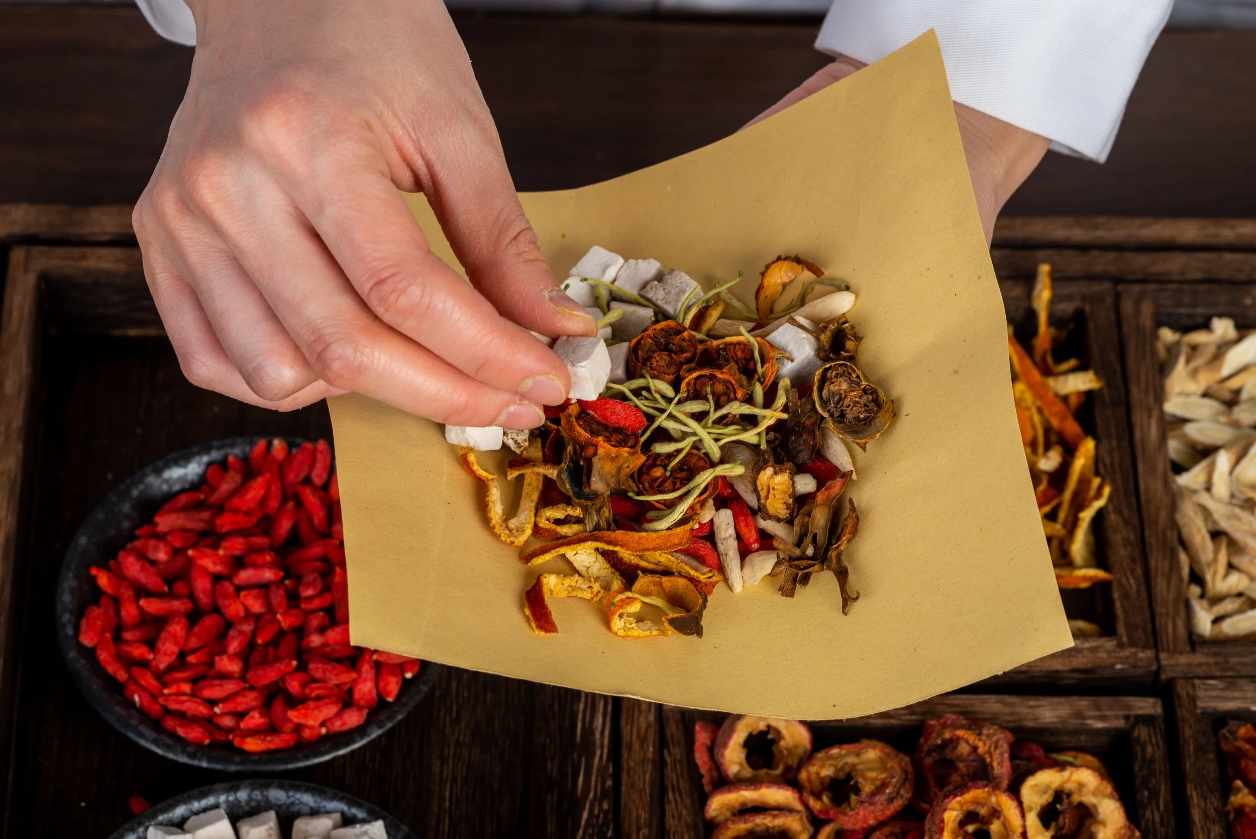 A hand placing dried herbs and flowers on a piece of parchment paper, with bowls of dried red berries and other dried herbs on a wooden surface.