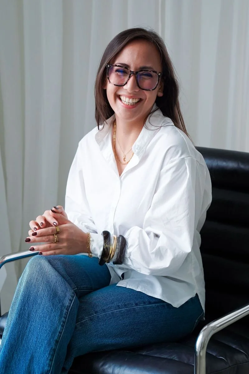 Eine lachende Frau mit langen braunen Haaren, Brille und weißen Hemd sitzt auf einem schwarzen Stuhl vor einem Vorhang.