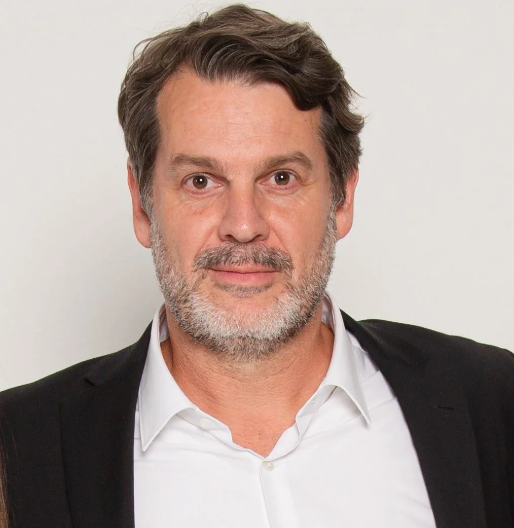 Ein Mann mit grauem Bart und braunem Haar, trägt einen schwarzen Blazer und ein weißes Hemd, auf weißem Hintergrund.