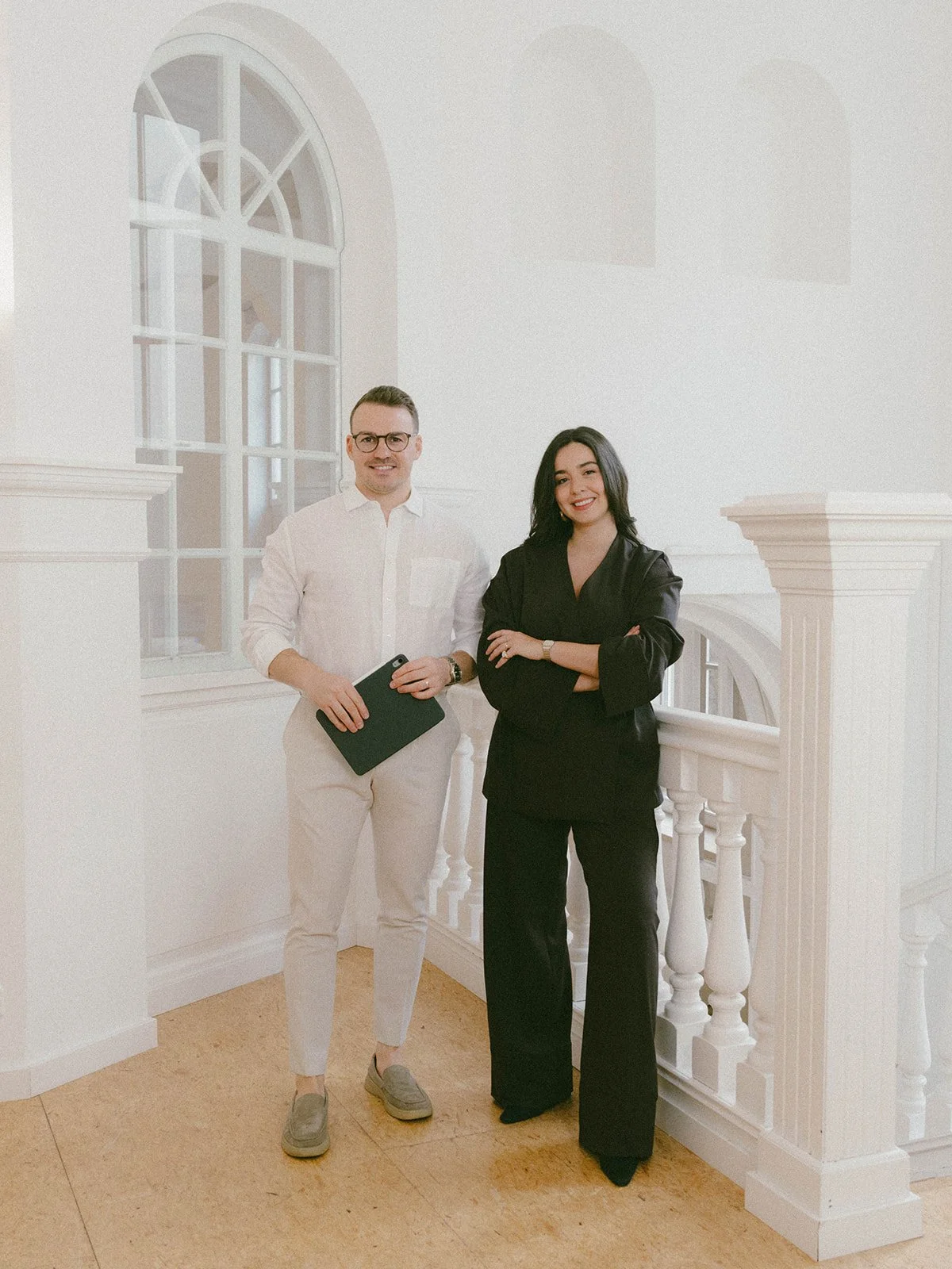 Zwei professionell gekleidete Personen, ein Mann und eine Frau, stehen an einer weißen, klassischen Treppe in einem hellen Raum mit großen Fenstern.