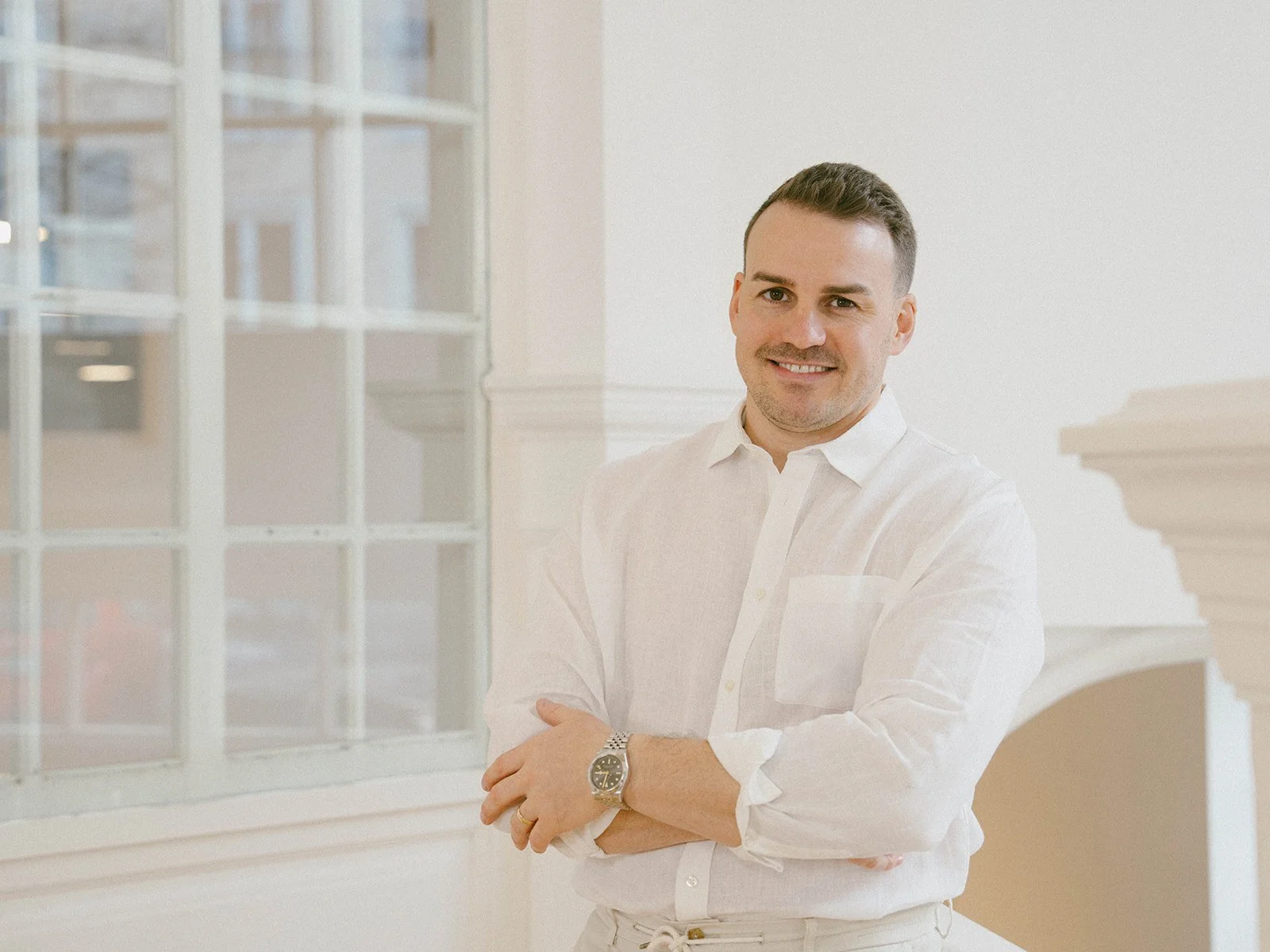 Ein Mann in weißem Hemd steht vor einem Fenster und lächelt in die Kamera.