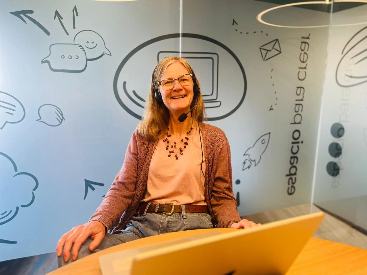 A woman is sitting in a room, smiling, a computer in front of her.