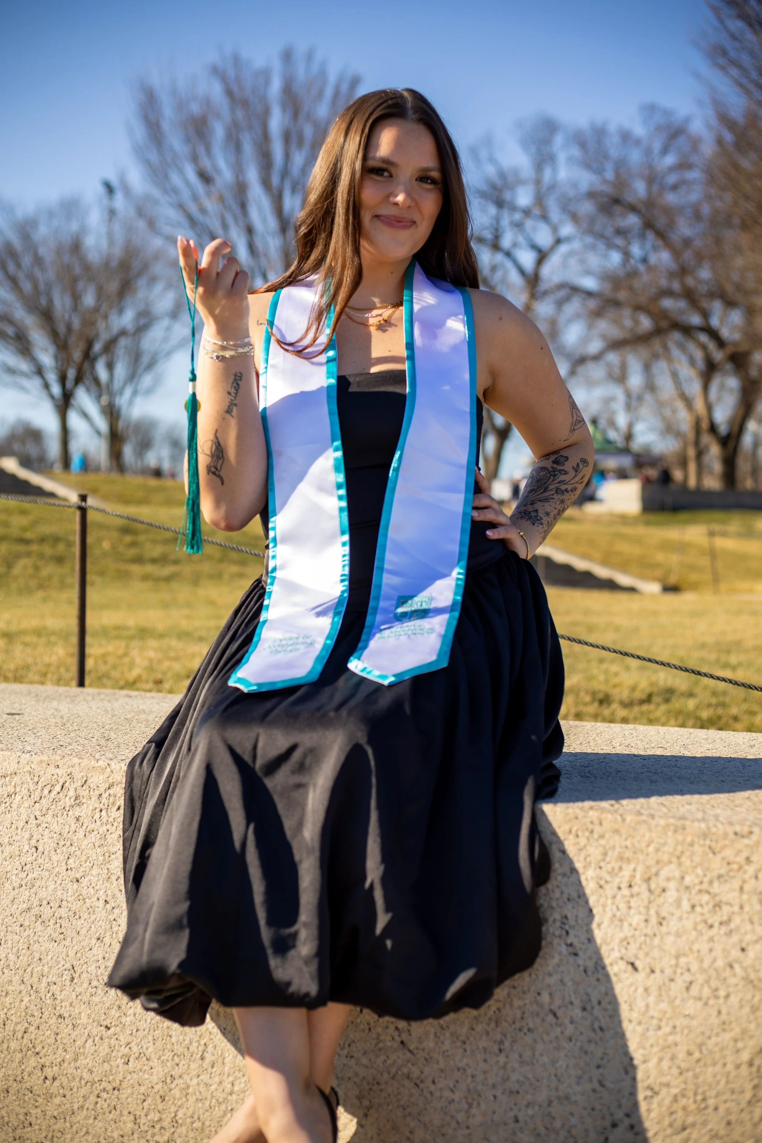 Young woman in a graduation gown sitting outdoors near a concrete ledge, smiling, with trees and a clear blue sky in the background.