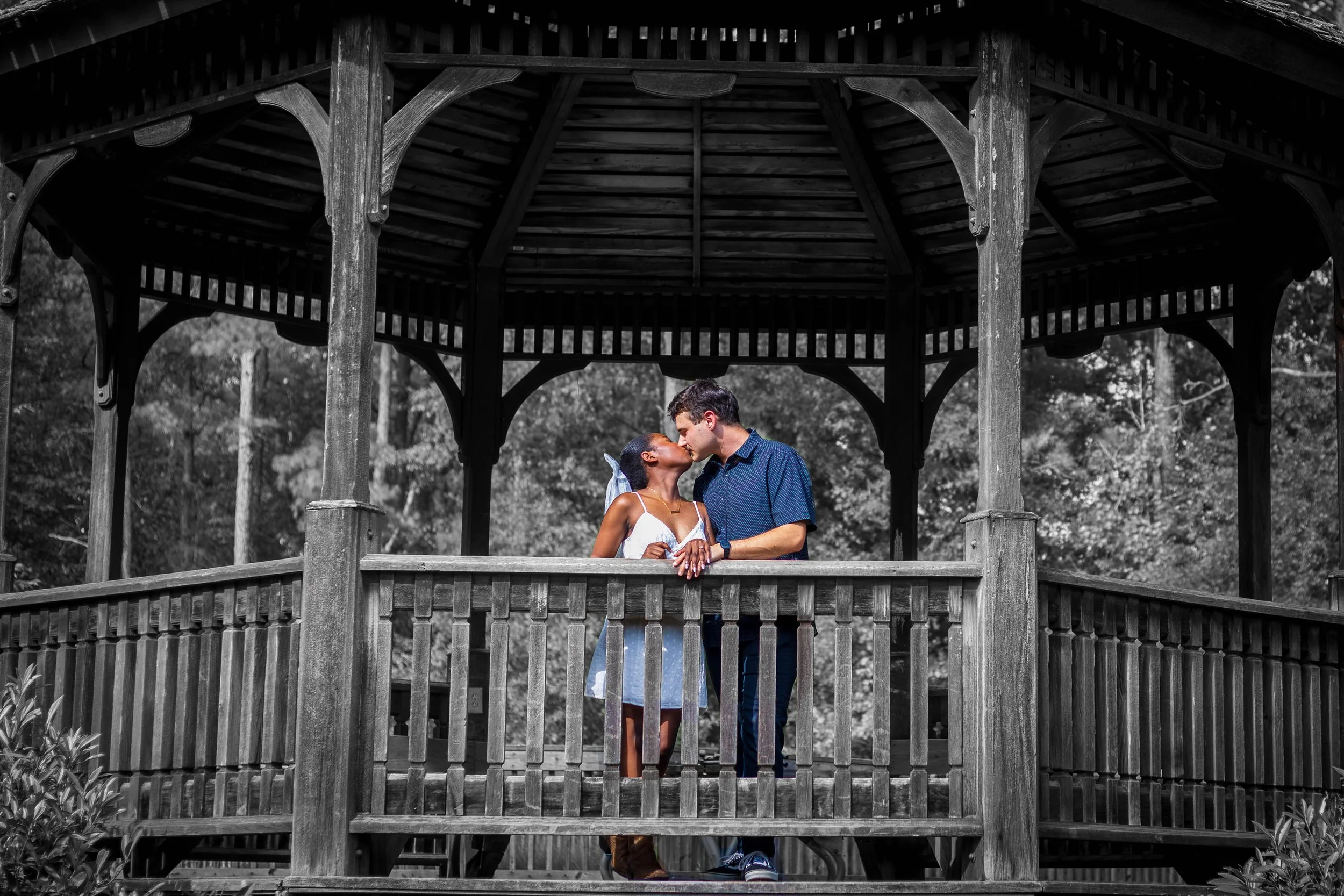 A couple sharing a kiss on a wooden gazebo, black and white background with trees.