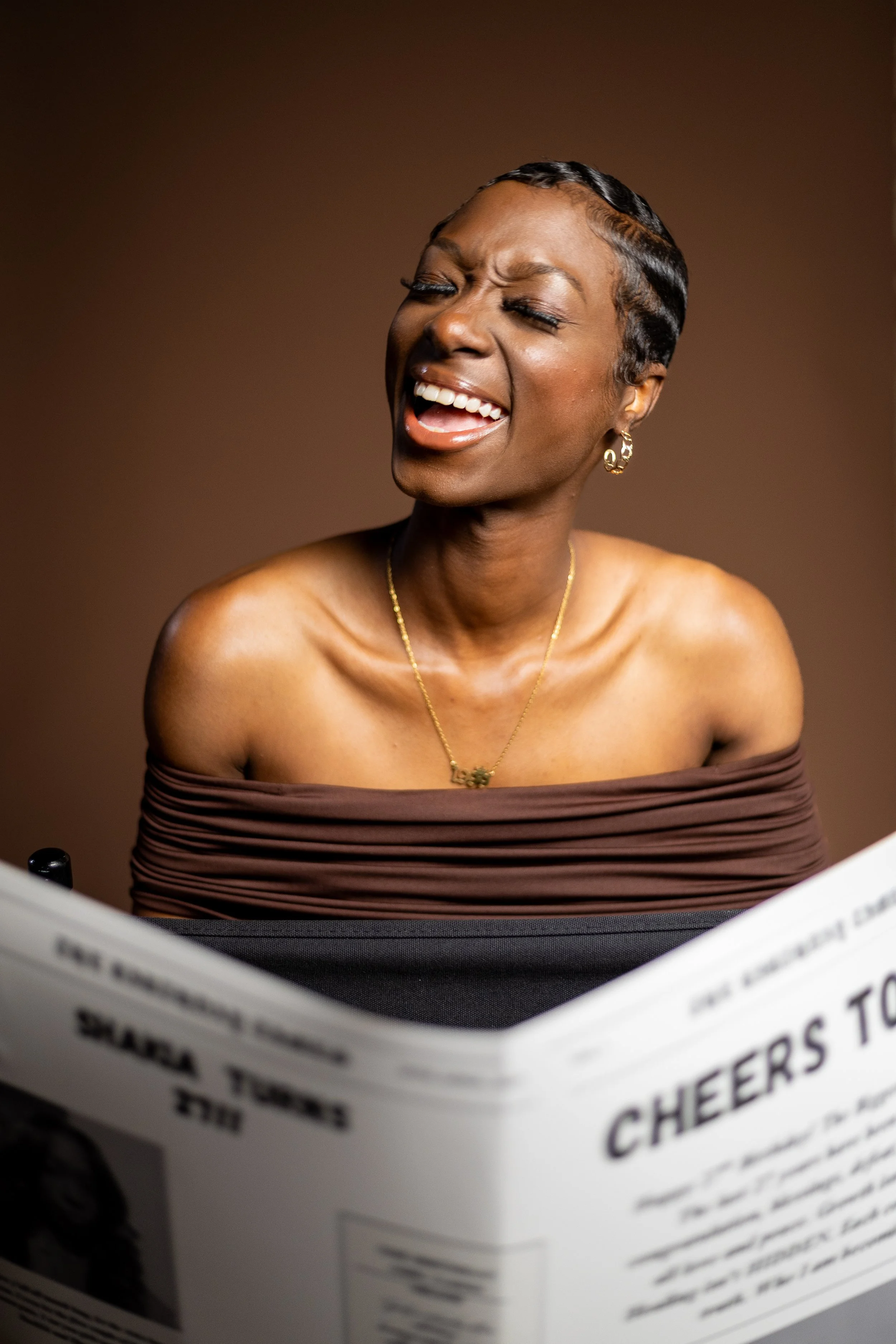 A woman with short black hair and gold earrings reading a newspaper and singing or speaking with her eyes closed, wearing an off-the-shoulder brown top against a brown background.