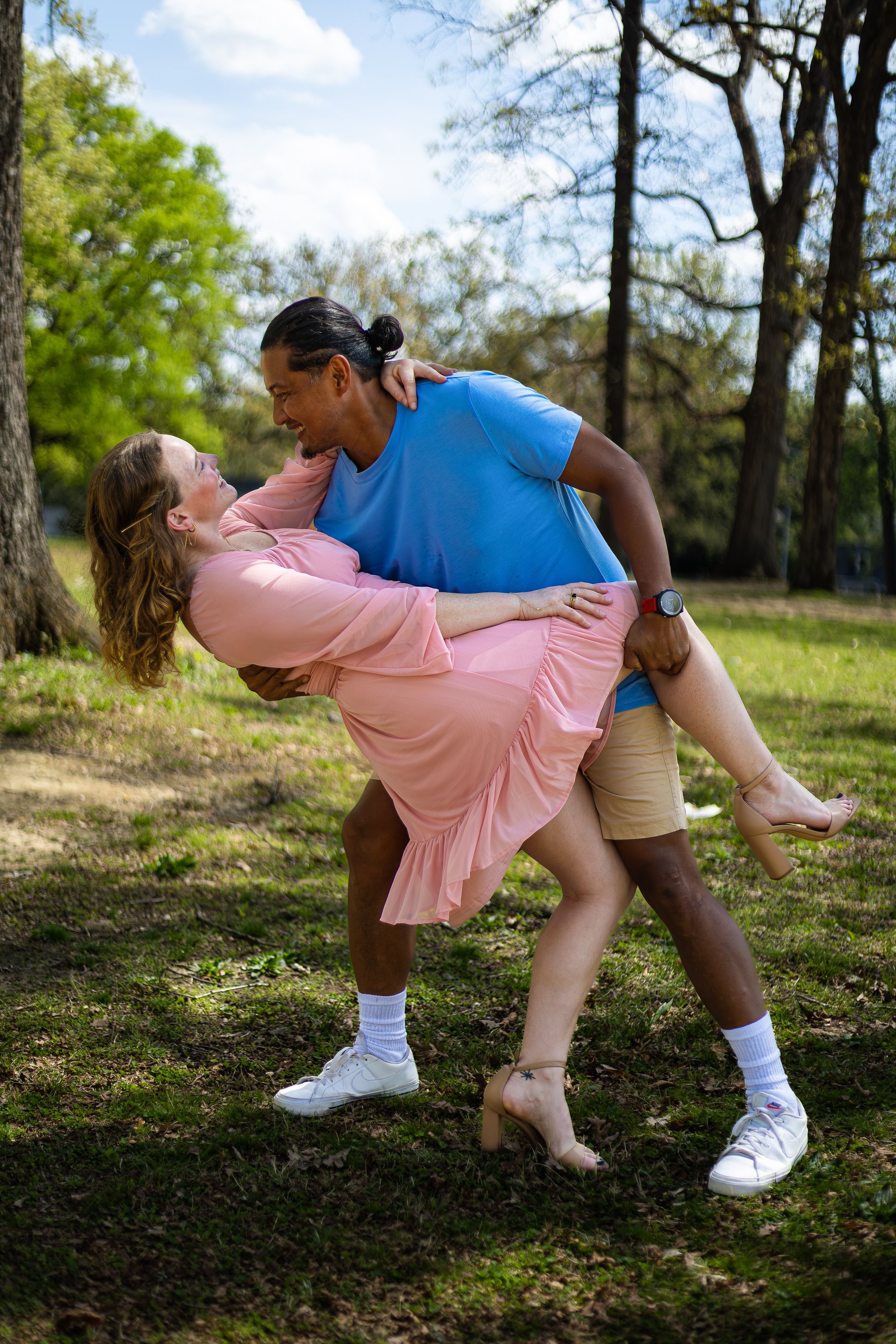 A man and woman dancing outdoors in a park with trees and grass, the man lifting the woman in a dip and both smiling.