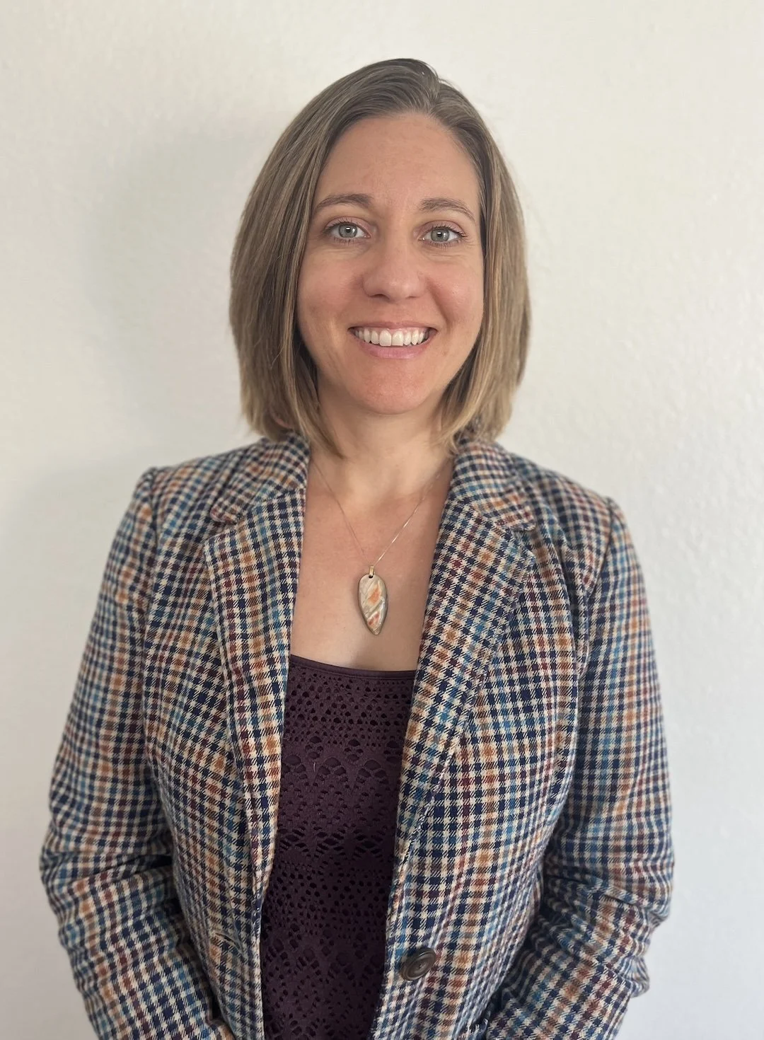 A woman with shoulder-length light brown hair, wearing a checkered blazer and a necklace with a stone pendant, standing against a plain white wall.