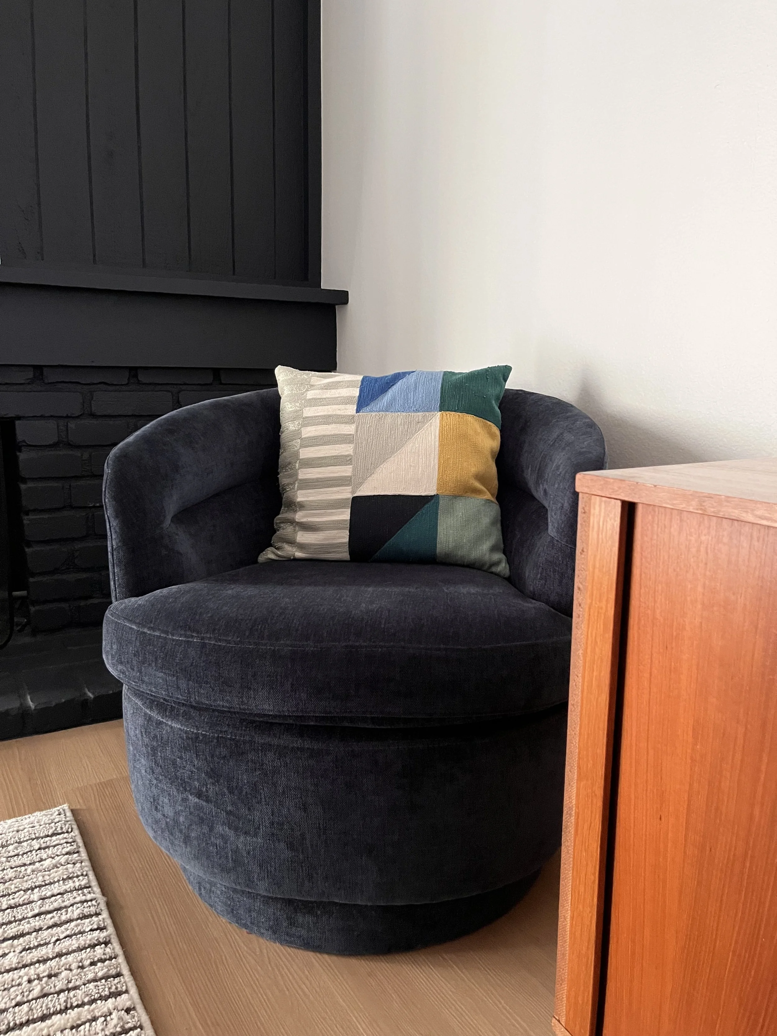 A dark gray velvet armchair with a colorful geometric pillow resting on it. The armchair is next to a wooden side table, with a black brick wall and a white wall in the background, and a small rug on the light wooden floor.