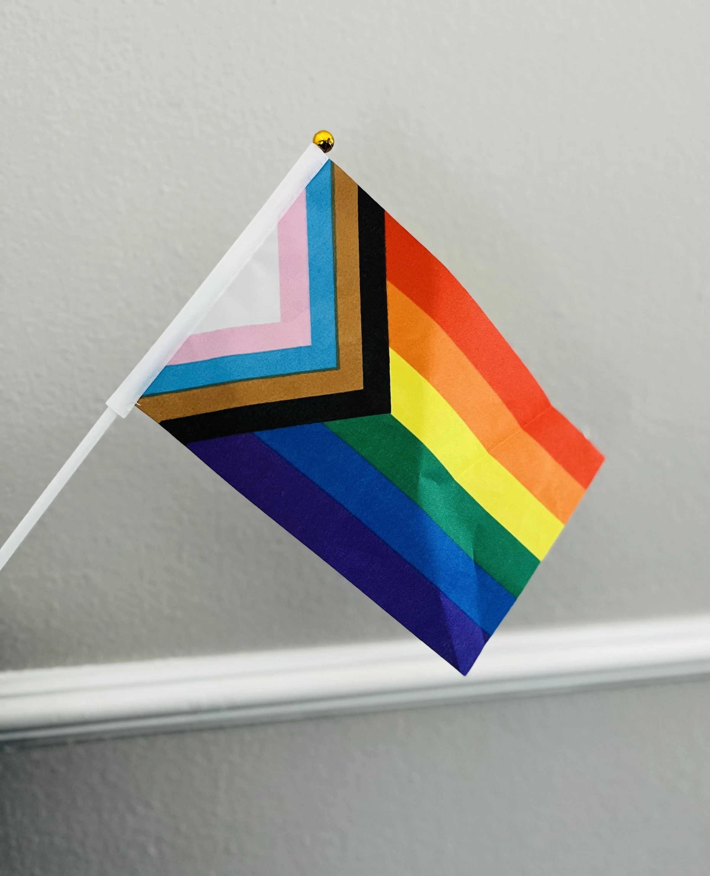 A small pride flag with rainbow colors is attached to a white stick. It is pinned to a white wall near a window or ledge, with a silver nail or pushpin at the top corner.