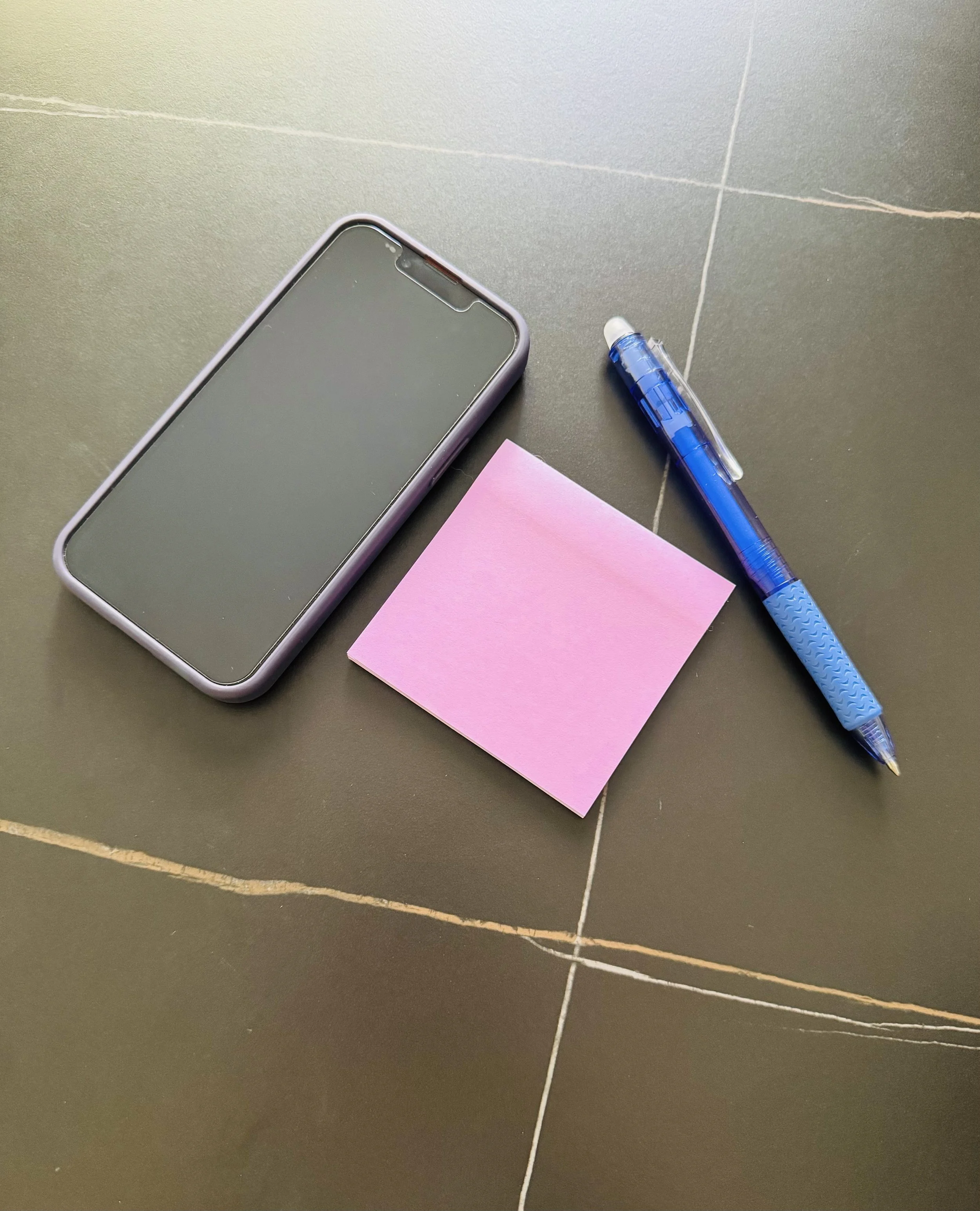 A smartphone with a gray case, a blue ballpoint pen, and a pink sticky note placed on a dark tiled surface.