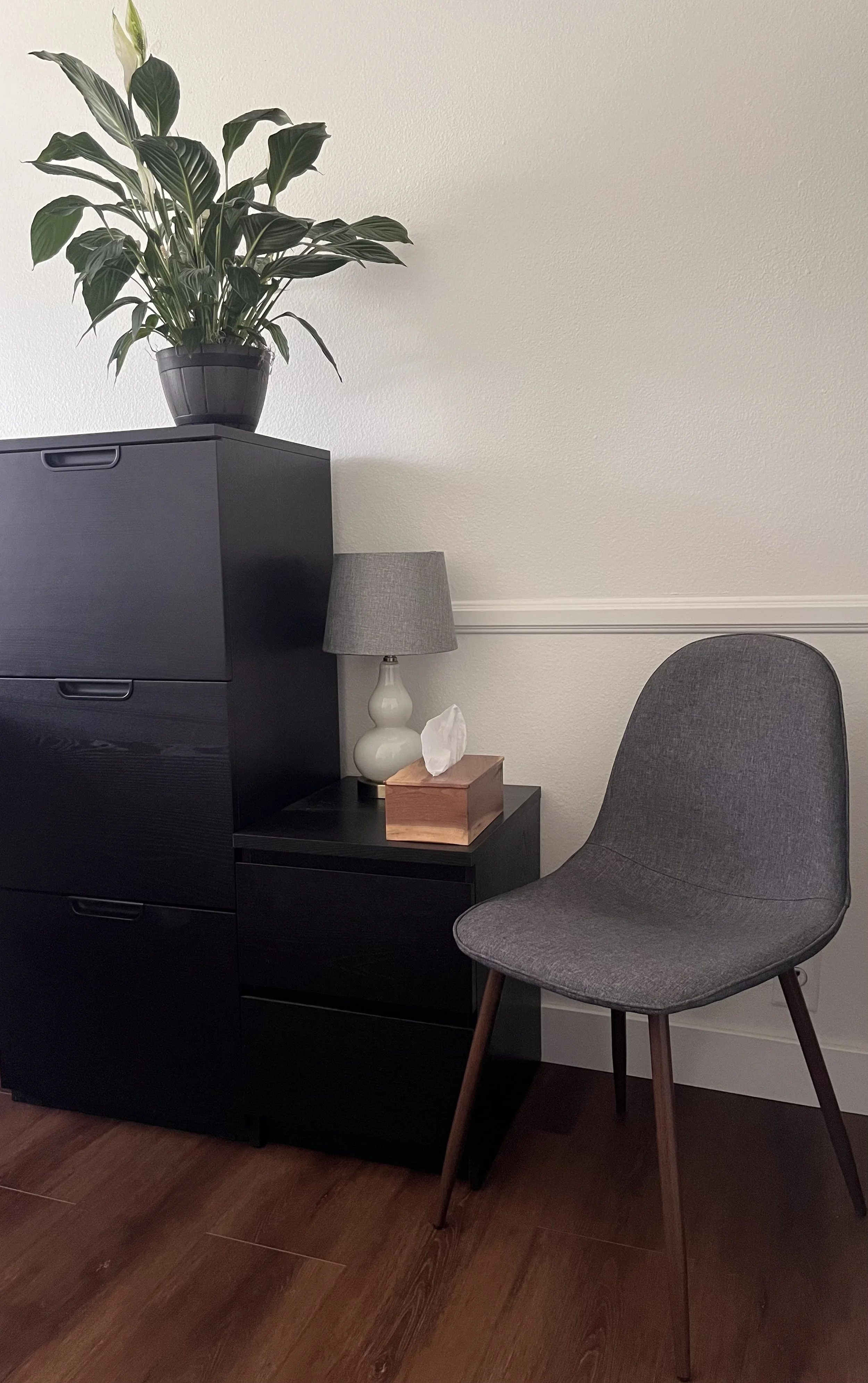 A small black cabinet with a potted plant on top, a lamp, and a tissue box on a side table next to a gray upholstered chair with wooden legs, set against a cream wall with white trim and brown hardwood floors.