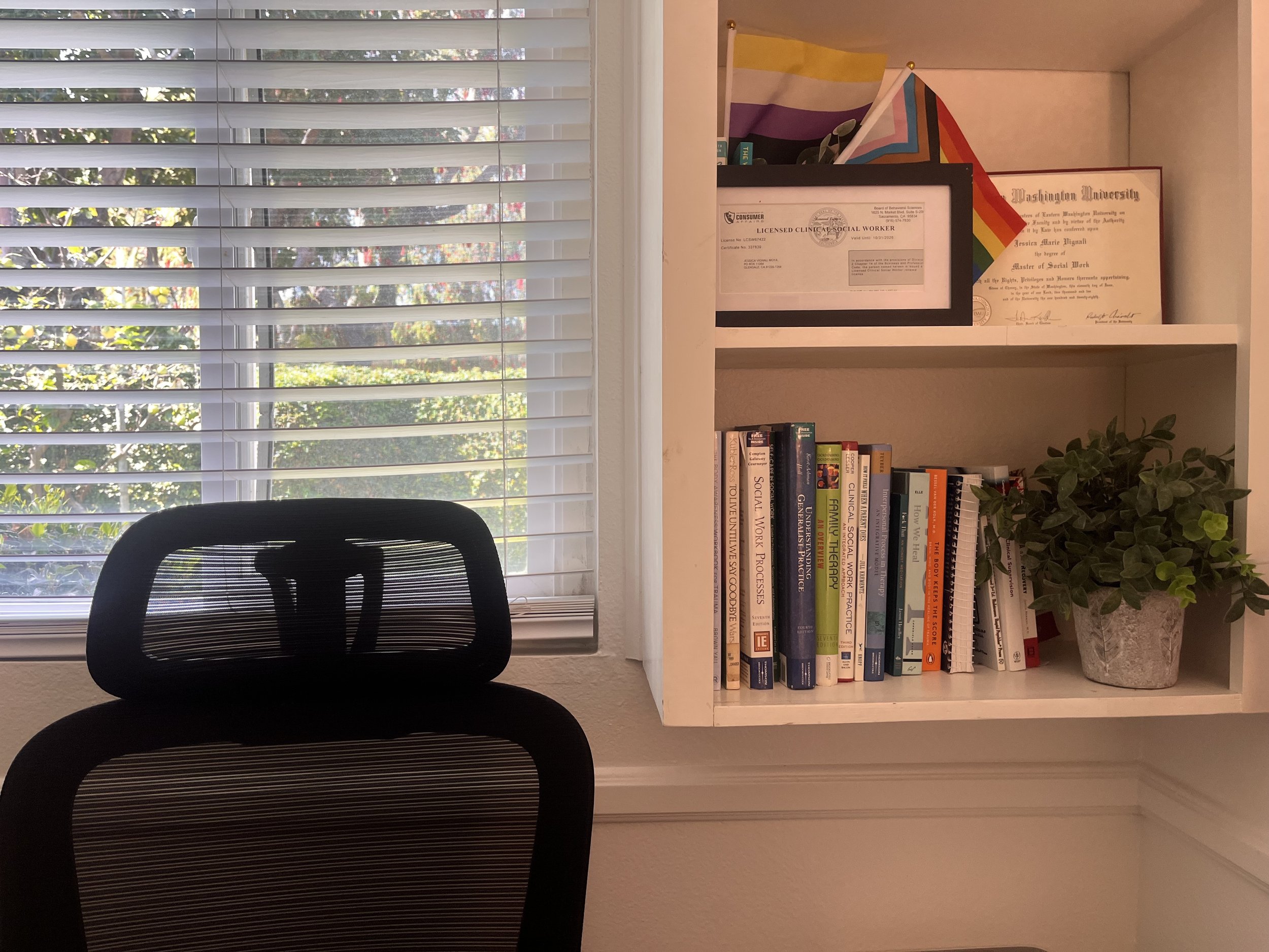 A black office chair in front of a white desk, with shelves containing books and a potted plant. Behind the setup, there is a window with white blinds and outside trees visible.