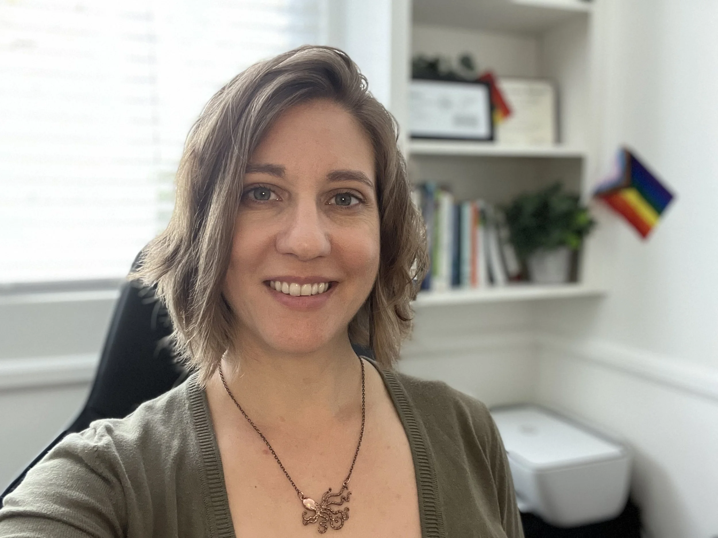 Close-up of a smiling woman with light brown hair, wearing a gray top and a decorative necklace, seated in an office environment with shelves, books, and a rainbow pride flag in the background.