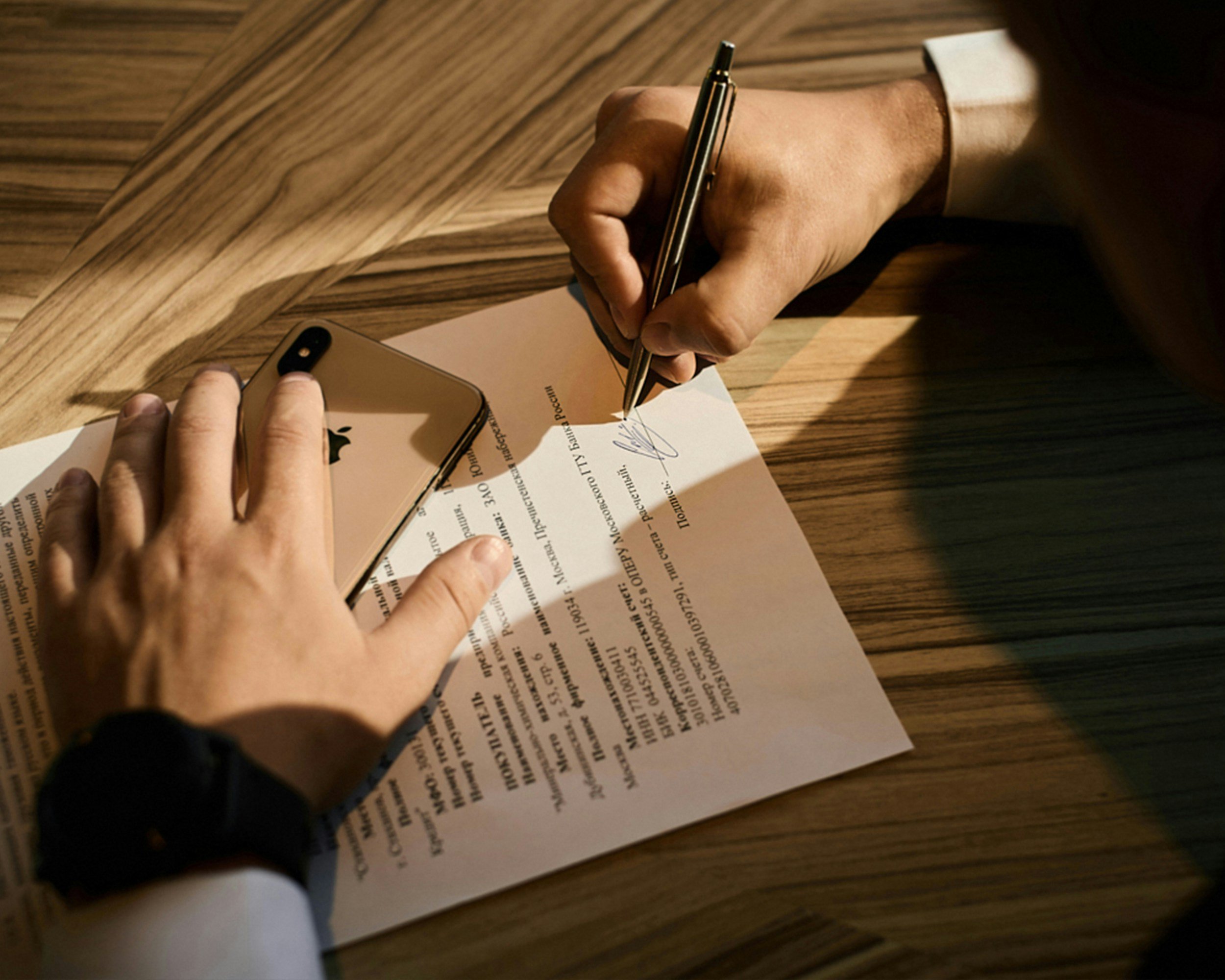 Someone signing a document on a wooden desk with an iPhone placed on the paper and a pen in hand.