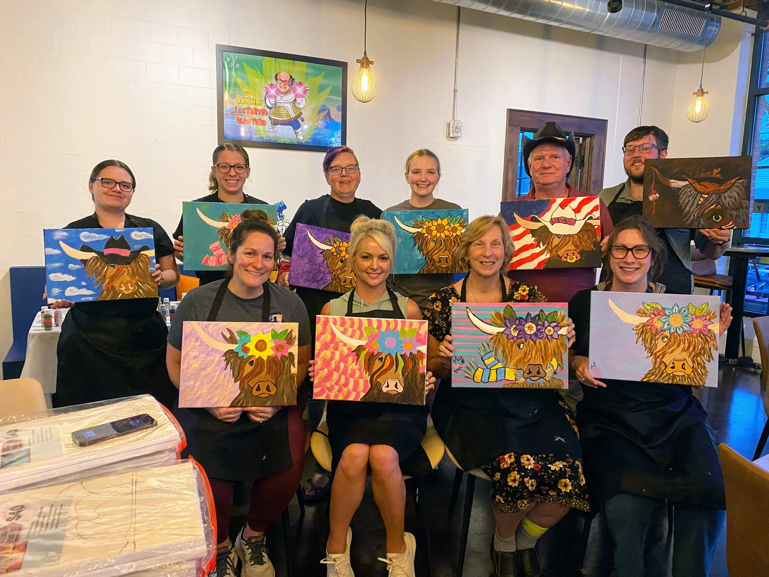 A group of people holding paintings of a highland cow with colorful floral and abstract backgrounds, posing indoors at an art event or class.