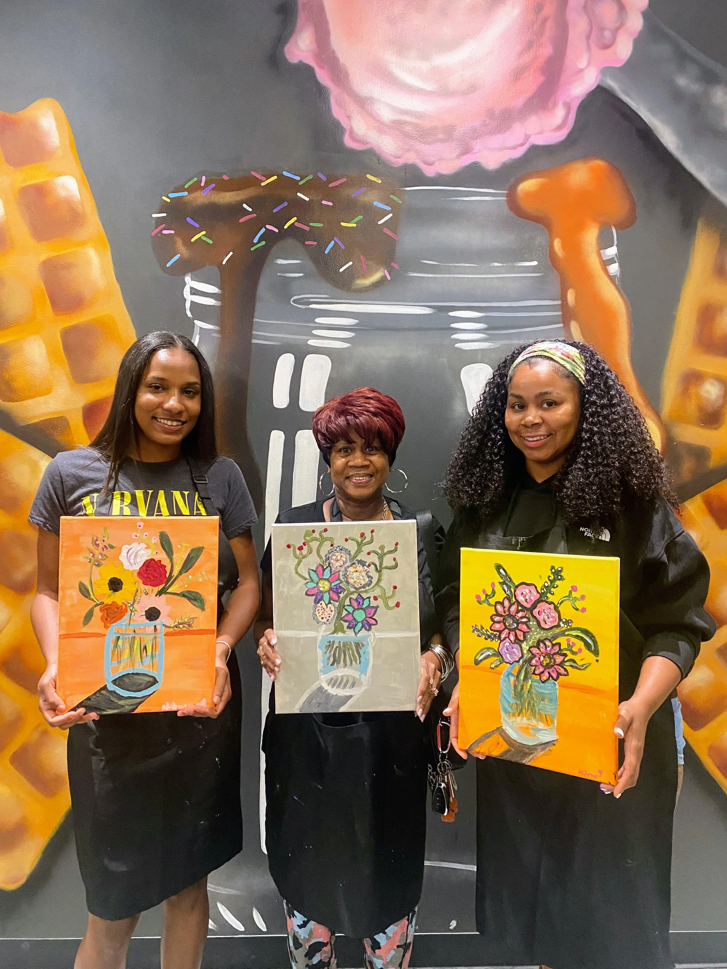 Three women standing in front of a large mural wall with artistic depictions of a mason jar, ice cream and sweets. Each woman is holding or displaying a colorful painting of a mason jar with flowers.