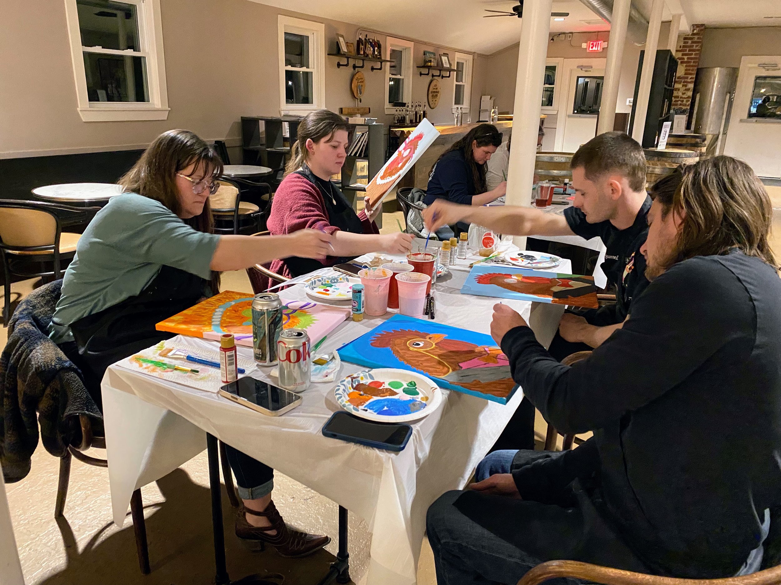 A group of people engaged in a painting activity around a table in a cozy indoor space, with paint supplies and artworks on the table.
