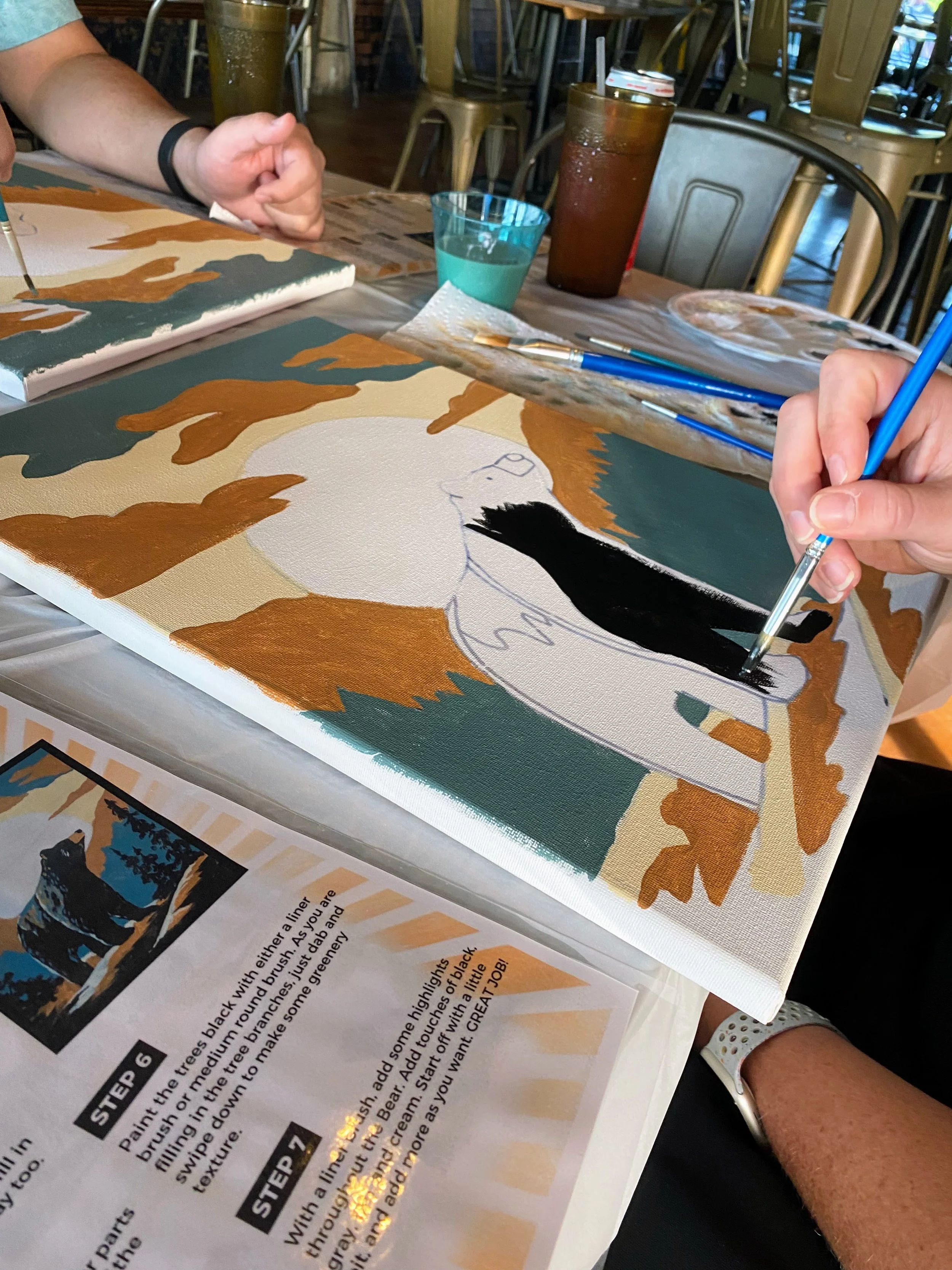 People painting a mountain scene with a bear on canvas at a table in a restaurant or studio space, with painting supplies and drinks nearby.