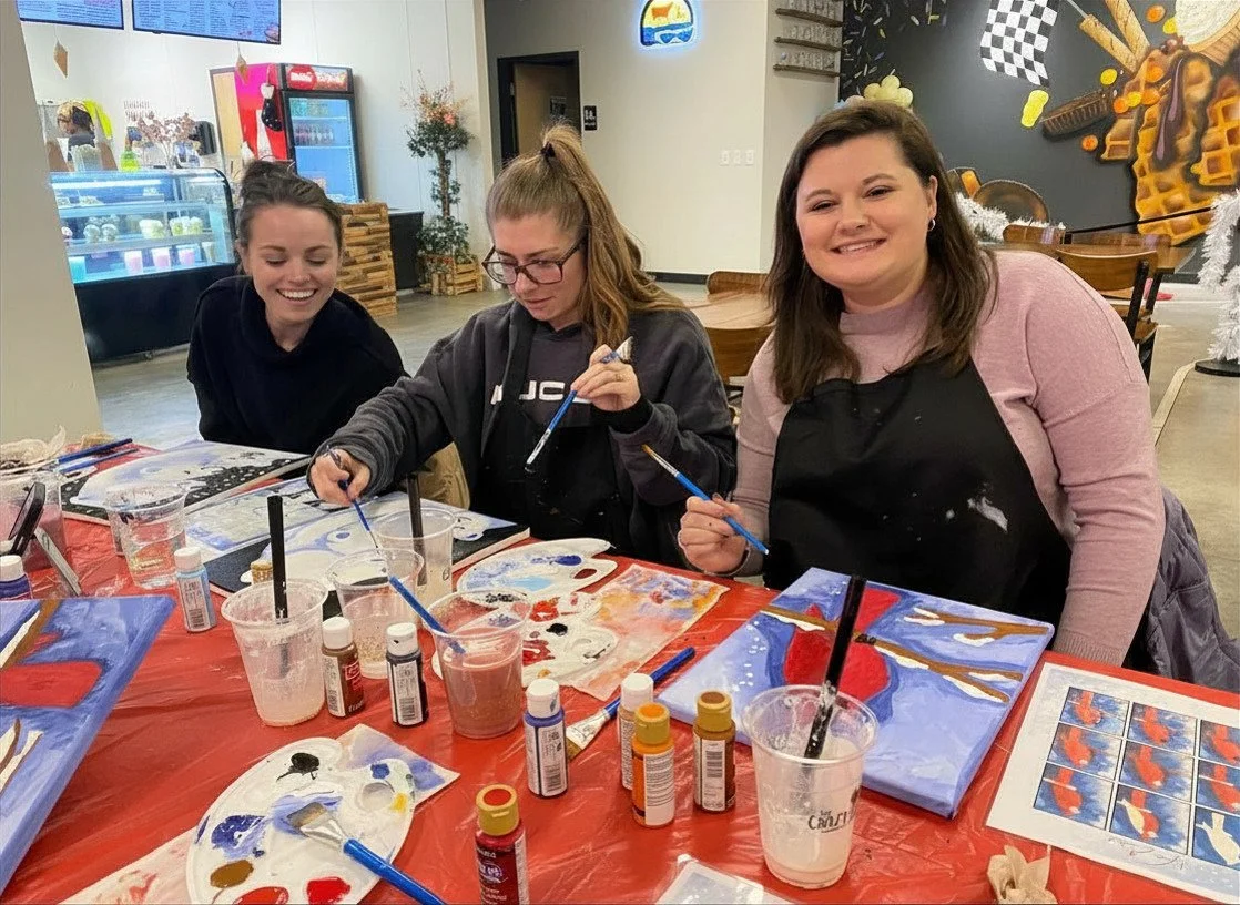 Three women are sitting at a table painting on canvases in a milkshake shop. They are smiling and appear to be enjoying a group painting activity with various paint bottles, brushes, and palettes on the table.