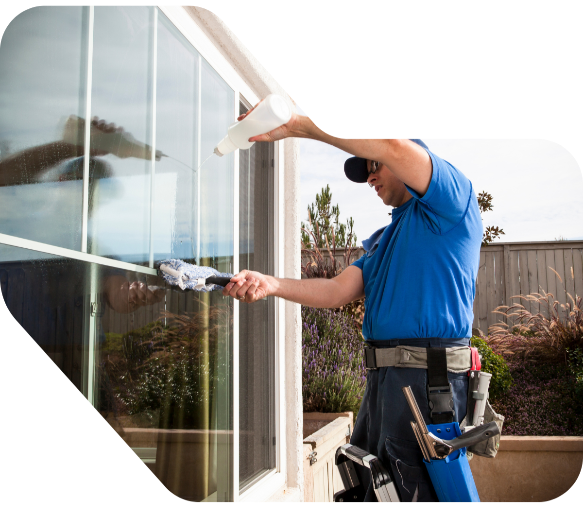 Un hombre limpiando una ventana de vidrio con una esponja y rociando con un recipiente spray, en un jardín con plantas y un cercado de madera.