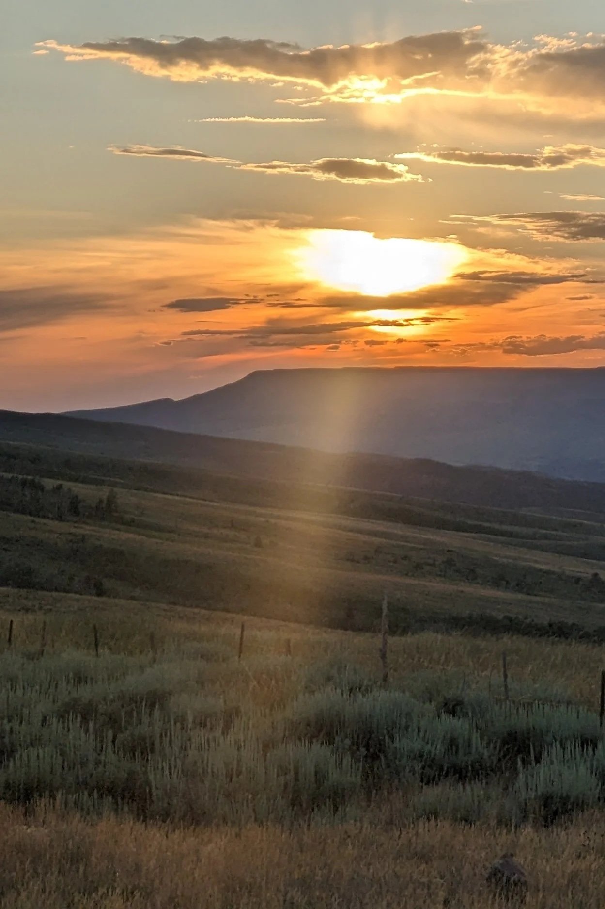 A landscape scene of rolling hills during sunset, with the sun partially hidden behind clouds, casting rays of light across the sky, and a distant mountain silhouette.