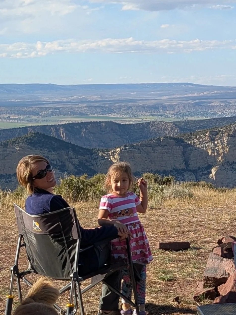 A woman and a young girl sitting and standing outdoors on a hill, with a vast mountainous landscape and distant plains under a partly cloudy sky in the background.