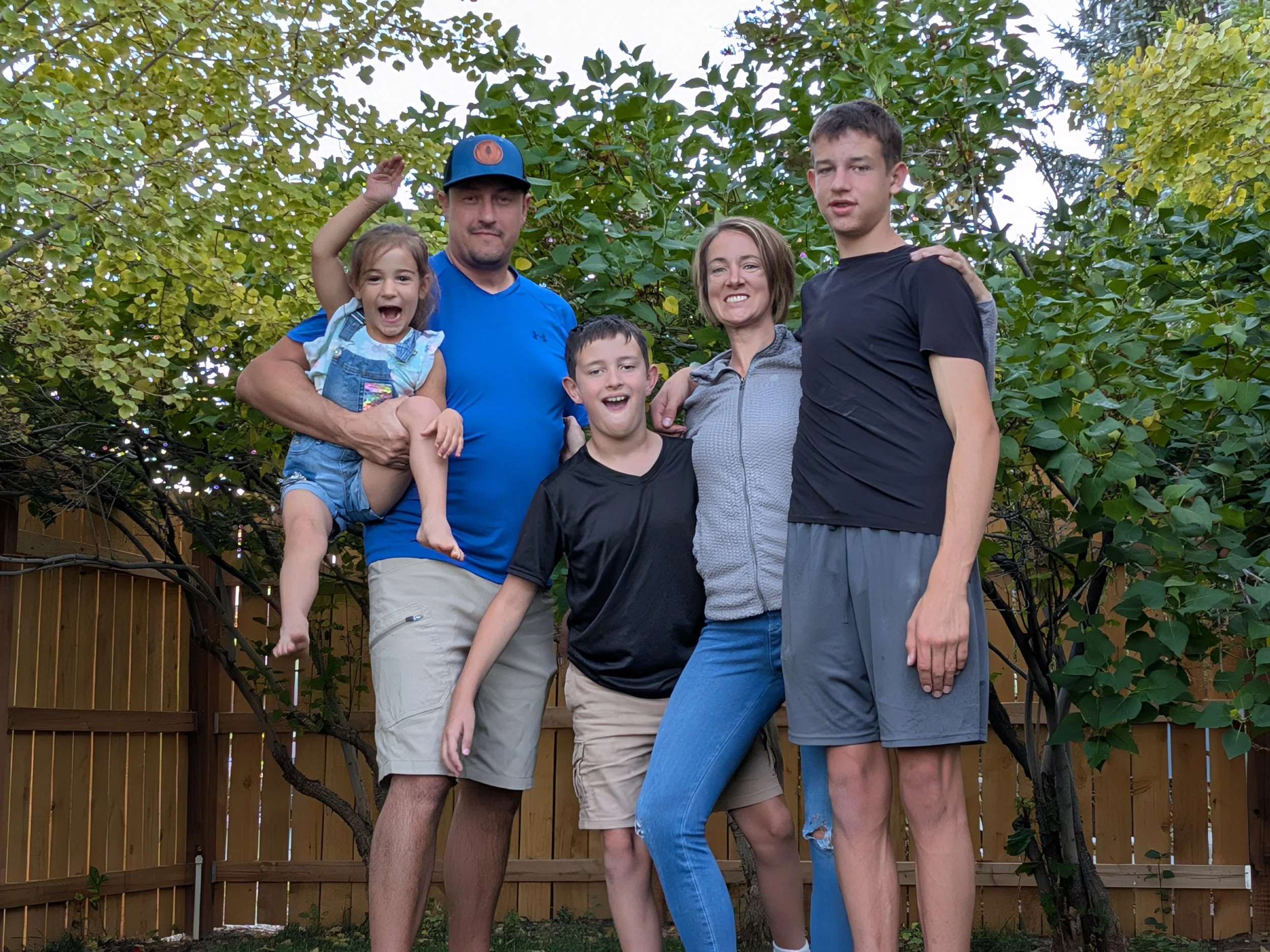 A group of six people, including two children, standing outdoors in front of green trees and a wooden fence, smiling and posing for a photo.