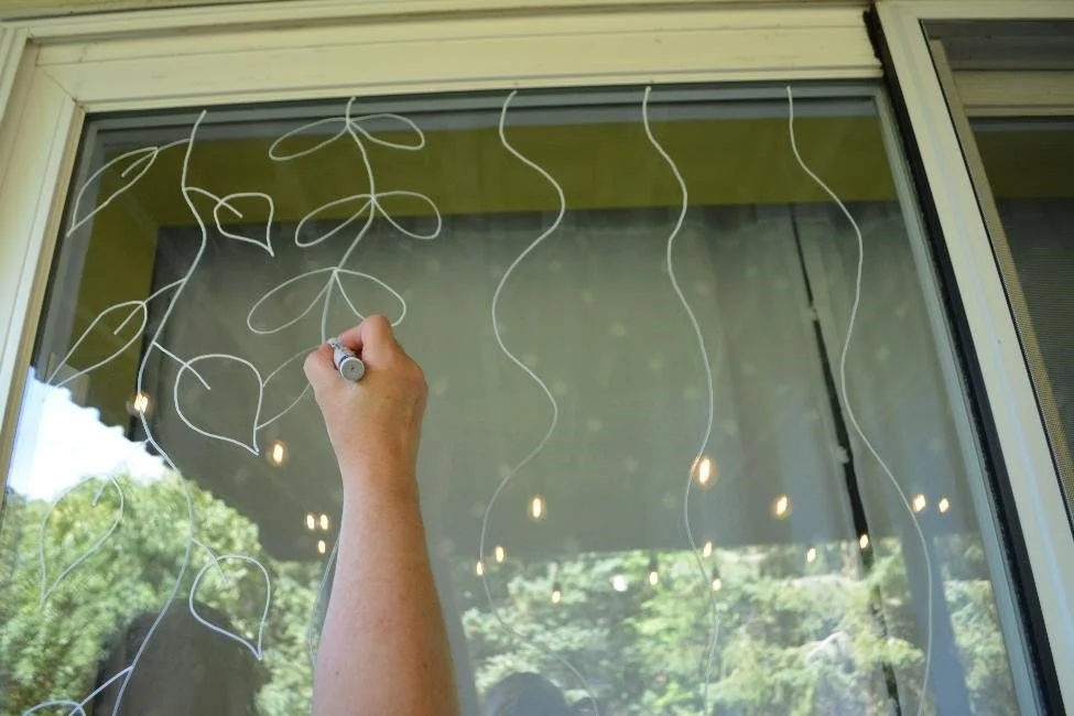 Person adding leaf designs onto wavy lines using oil paint marker on exterior window to create bird-safe window solution