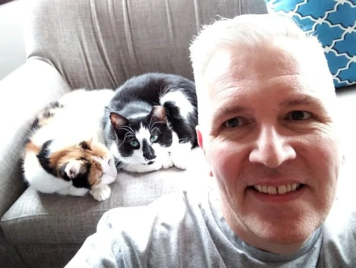 Photo of Paul taking a selfie at home with his two calico cats sitting behind him on a couch