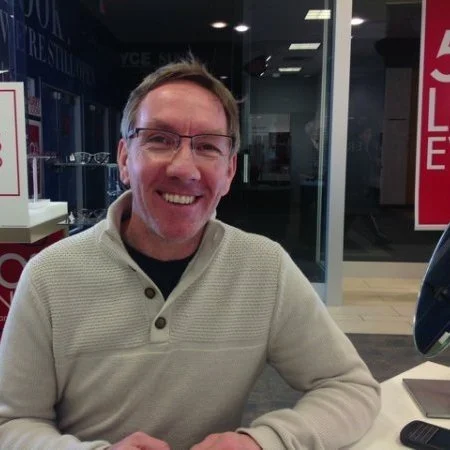 Photograph of smiling Brian Finlayson in a warm sweater and glasses smiling straight at the camera while seated by a desk instead of an office