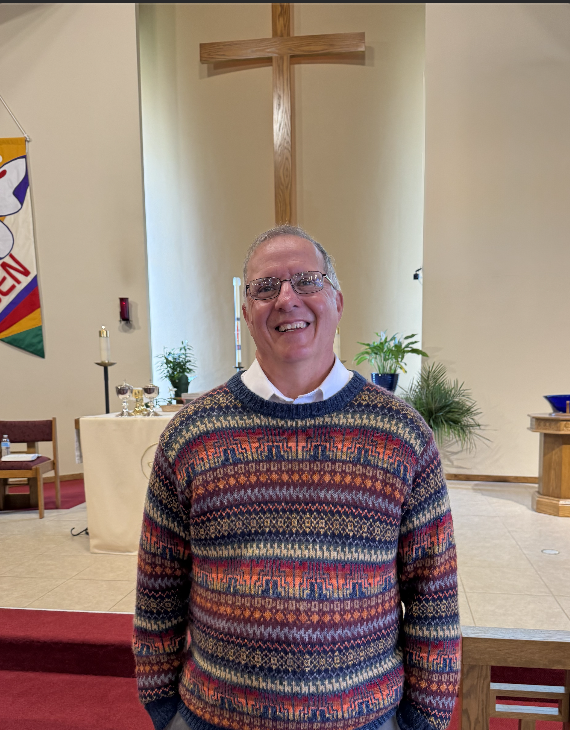 Man smiling inside a church, standing in front of an altar with a wooden cross behind him, decorated with candles and plants.