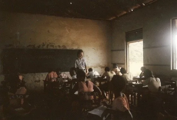 A classroom with students sitting at desks and a teacher standing at the front near a blackboard, in a poorly lit room with natural light coming through an open window.