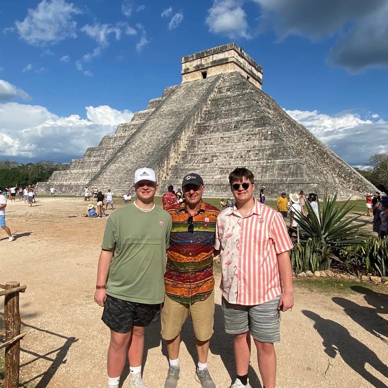 Three young men standing in front of the pyramid of Kukulkan at Chichen Itza, under a partly cloudy sky, with other tourists in the background.