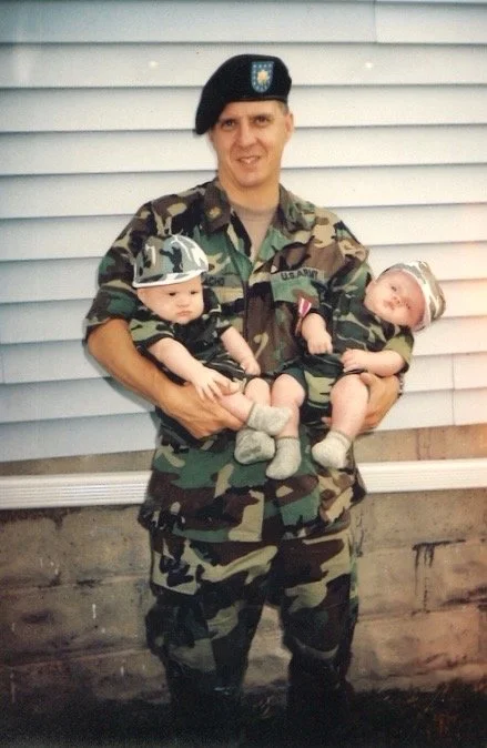 A man in U.S. Army camouflage uniform holding two young children, all wearing matching camouflage outfits, in front of a white siding house.